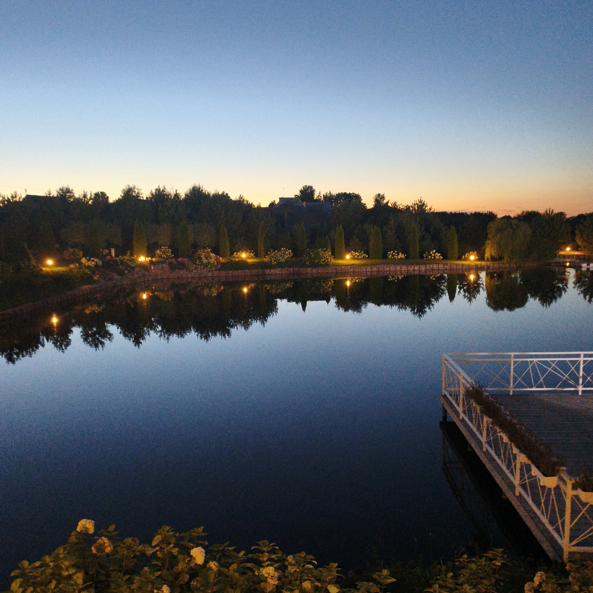 This is a photograph of a lake at dusk in Sapotskinski Sel'ski Sovet, Belarus. The sky is a gradient of deep blue at the top to a pale orange near the horizon, indicating the sunset is fading. The lake is calm and reflects the trees and lights along its bank. On the right side of the image, there is a white wooden pier with decorative railings, extending into the water. Along the shoreline, there are trees, bushes, and lights illuminating the landscape. Some flowers are visible in the foreground. There are no people present in the scene.