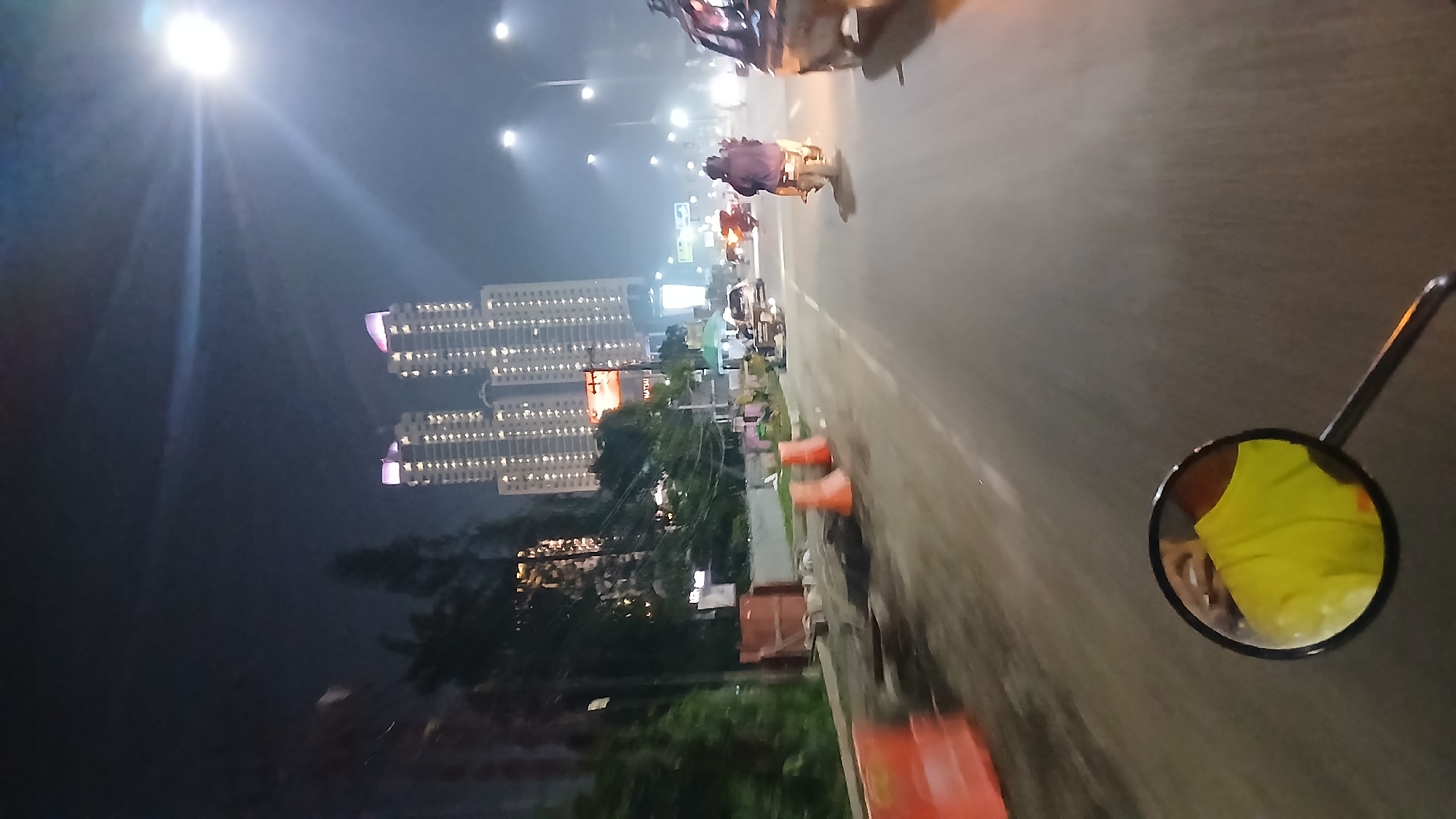 The image depicts a night-time street scene in the City of Medan, Indonesia. Two tall, modern apartment buildings with illuminated windows dominate the background. In the foreground, a road is visible, with a motorcycle in motion, seen from the perspective of its rider. The rider's bright yellow shirt is reflected in the motorcycle's side mirror. Traffic cones and construction barriers are present along the side of the road, suggesting ongoing road work. Several other vehicles, including another motorcycle and a car, are also on the road. Streetlights illuminate the scene, creating a somewhat hazy atmosphere.