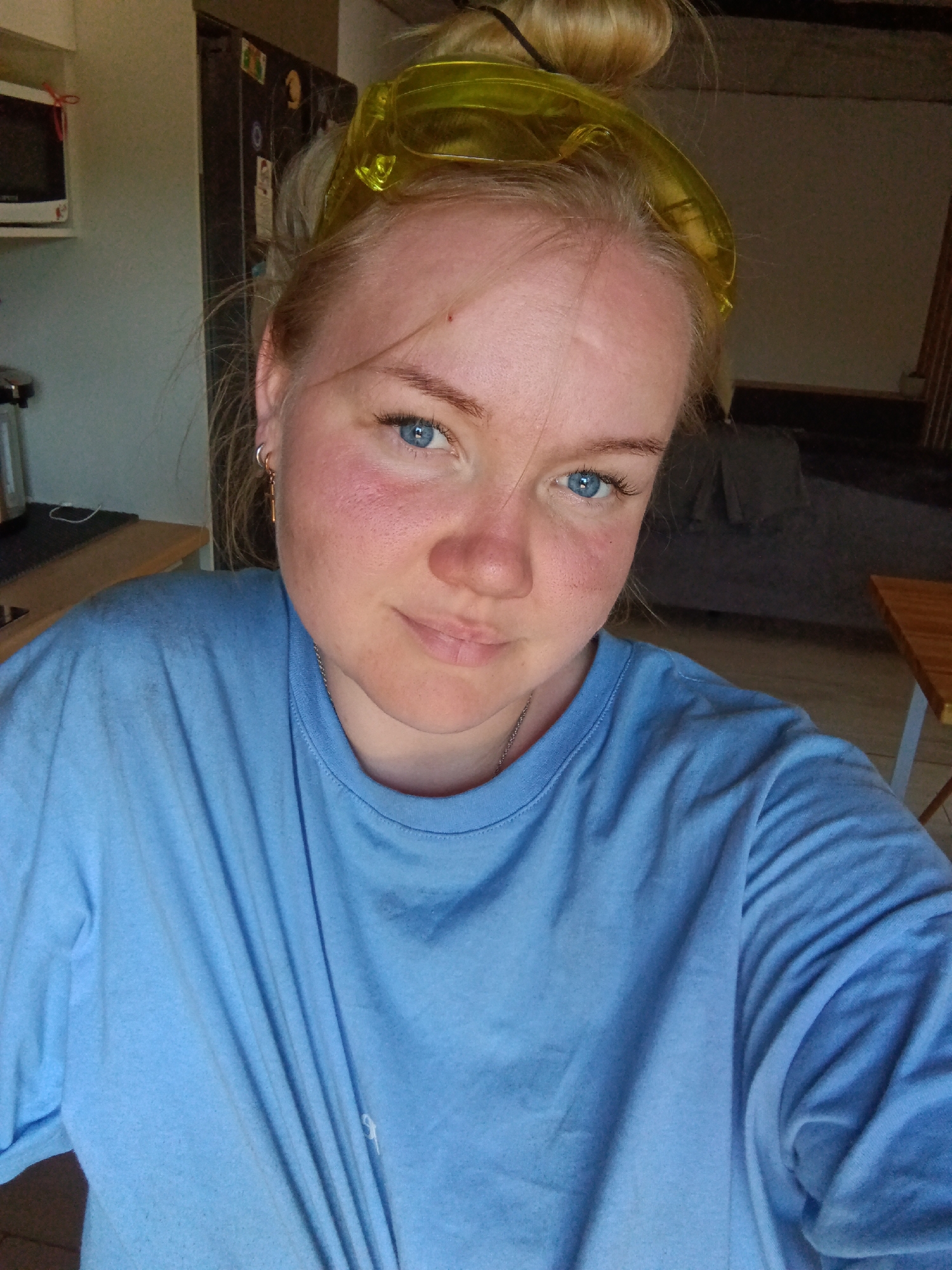 A fair-skinned woman with blonde hair pulled into a bun and wearing yellow goggles on her head is posing for a selfie indoors. She is wearing a light blue t-shirt. The background is a domestic kitchen setting with visible appliances and furniture. There is no indication of the location being in the city of Фоки, Russia from the image.