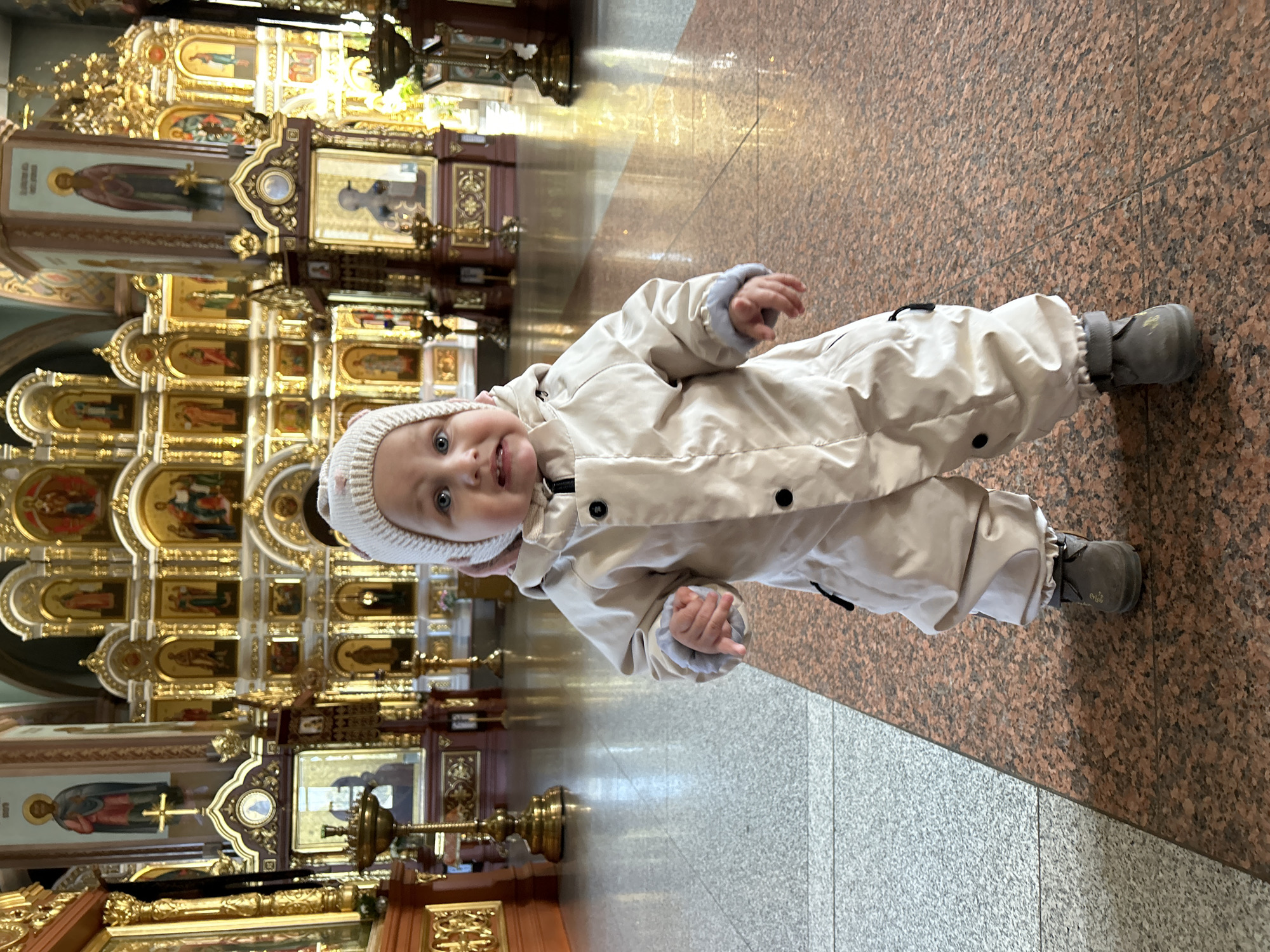 A young child wearing a cream-colored snowsuit and knit hat stands in a religious building in Chaikovsky, Russia. The building's interior has a marble-like floor of two distinct colors, and an ornate, multi-tiered, golden altar or iconostasis is visible behind the child.