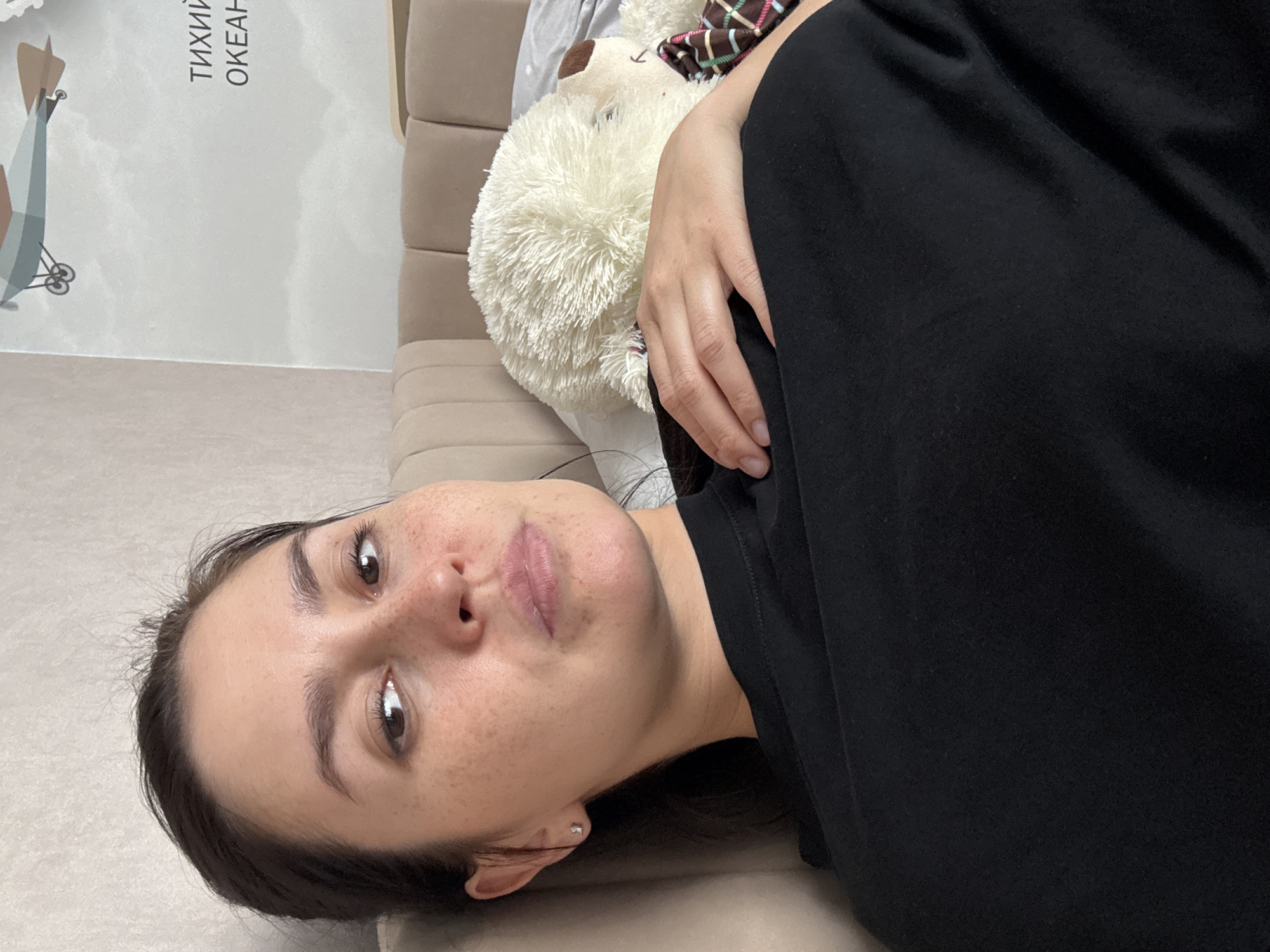 A young woman with dark hair and freckles is lying down, holding a large, fluffy white teddy bear. She is wearing a black t-shirt. The background shows a wall with a poster of an airplane and text in Russian. The room appears to be a bedroom, with a beige headboard visible. There is no indication of the city of Нижнеяркеево, Russia.