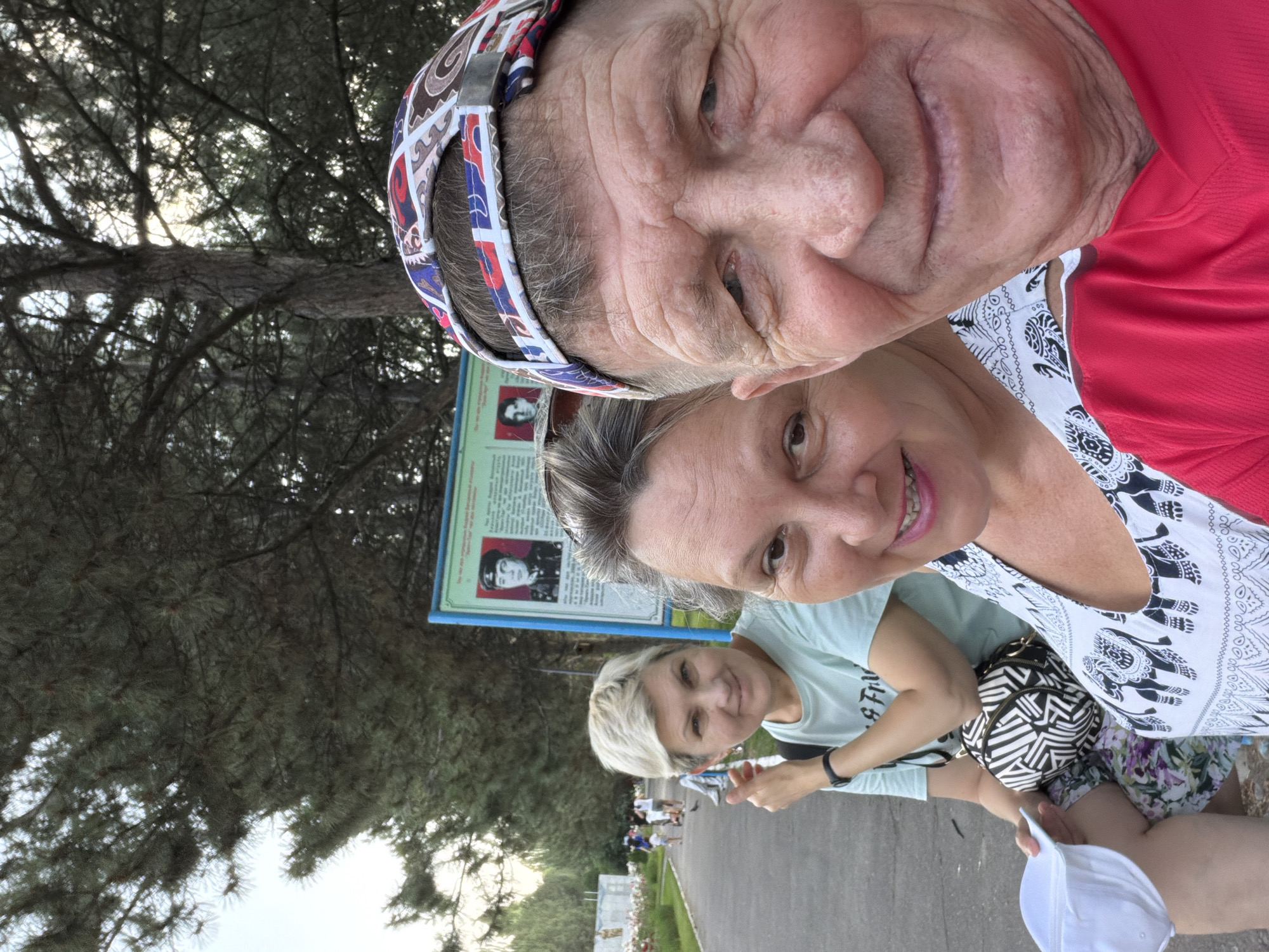 Three people are posing for a selfie in an outdoor park. In the foreground, a man wearing a red shirt and a colorful baseball cap is on the right, and a woman wearing a white and black patterned top is to his left. In the background, a woman with short blonde hair wearing a light blue t-shirt is also visible. She is sitting on a bench and holding a white hat. Behind them, there is a park pathway with trees lining the sides, and a display board with portraits and text. The lighting suggests it is daytime, possibly late afternoon.