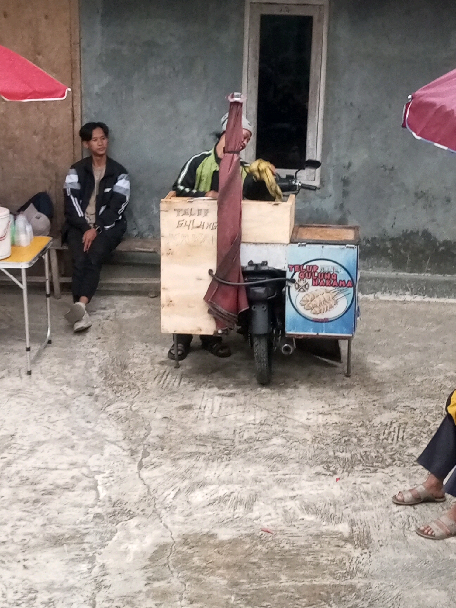 In Kalibeber, Indonesia, a young man with short dark hair is sitting on a wooden bench to the left of the frame. He is wearing a black jacket with white and gray stripes on the sleeves, a light-colored t-shirt, and black pants. Beside him is a small foldable yellow table with some white containers and bags on it.
To the right, a food cart is attached to a motorcycle. The cart is primarily made of light-colored wood with a blue advertisement panel on its side. The advertisement features an image of three skewers of food and text in Indonesian. A person wearing a black and green jacket is partially visible behind the food cart, appearing to be attending to it. A maroon umbrella is positioned over the motorcycle and cart.
The ground is a rough, textured concrete surface, with some debris scattered on it, including small red objects. The background consists of a plain, textured wall, with a vertical window set into it. To the far right, a person's legs and feet are visible, wearing dark pants and sandals. The scene suggests an outdoor, informal marketplace or roadside vendor setup.