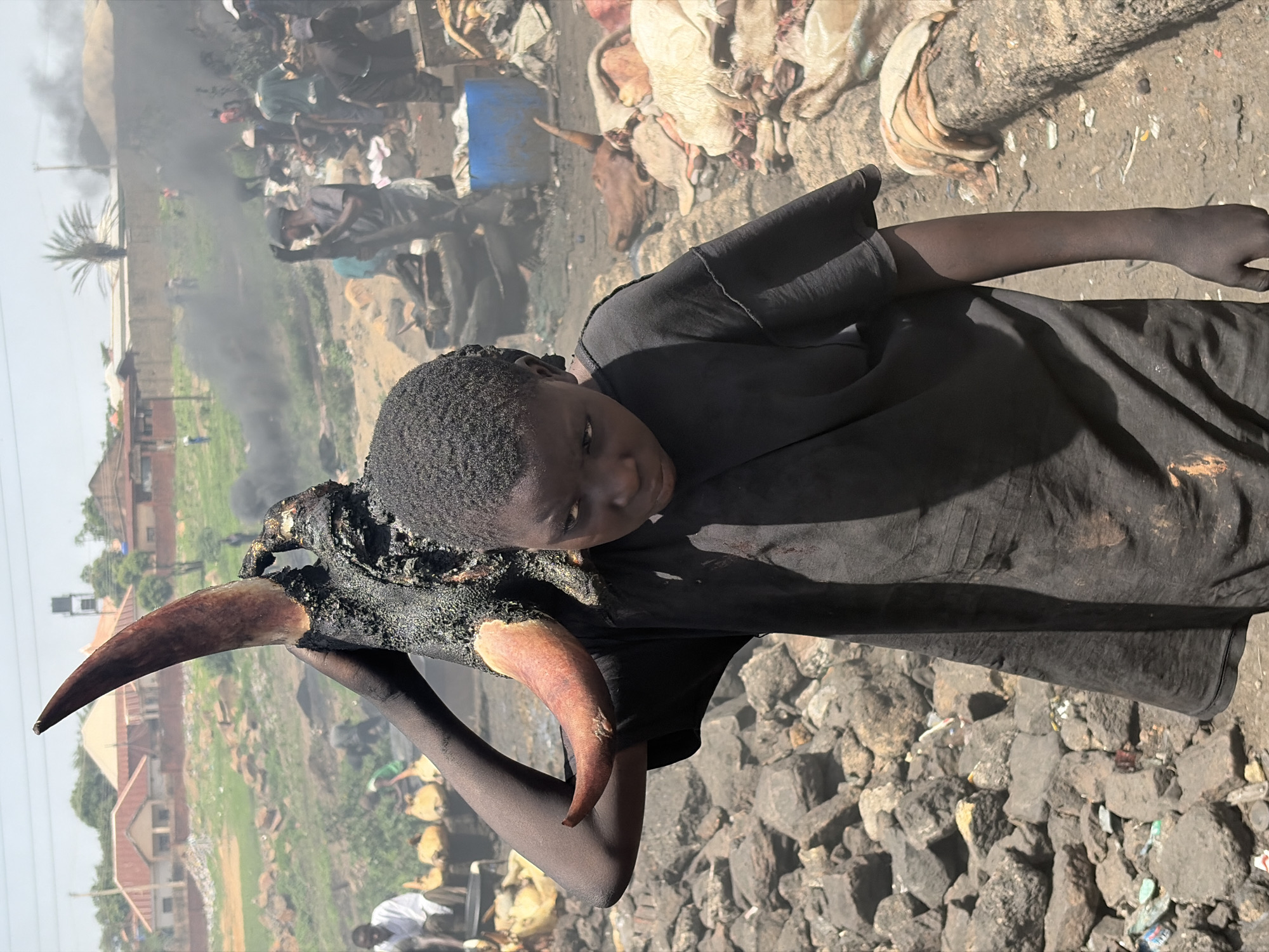 In Jos, Nigeria, a young person with dark skin and short hair, wearing a black t-shirt, holds a charred bull's head with long horns. The person's arms and hands appear blackened. The foreground shows rocky terrain. In the background, there are buildings, a palm tree, smoke, and several indistinct figures. The scene appears to be an outdoor area where animal hides and other materials are scattered.
