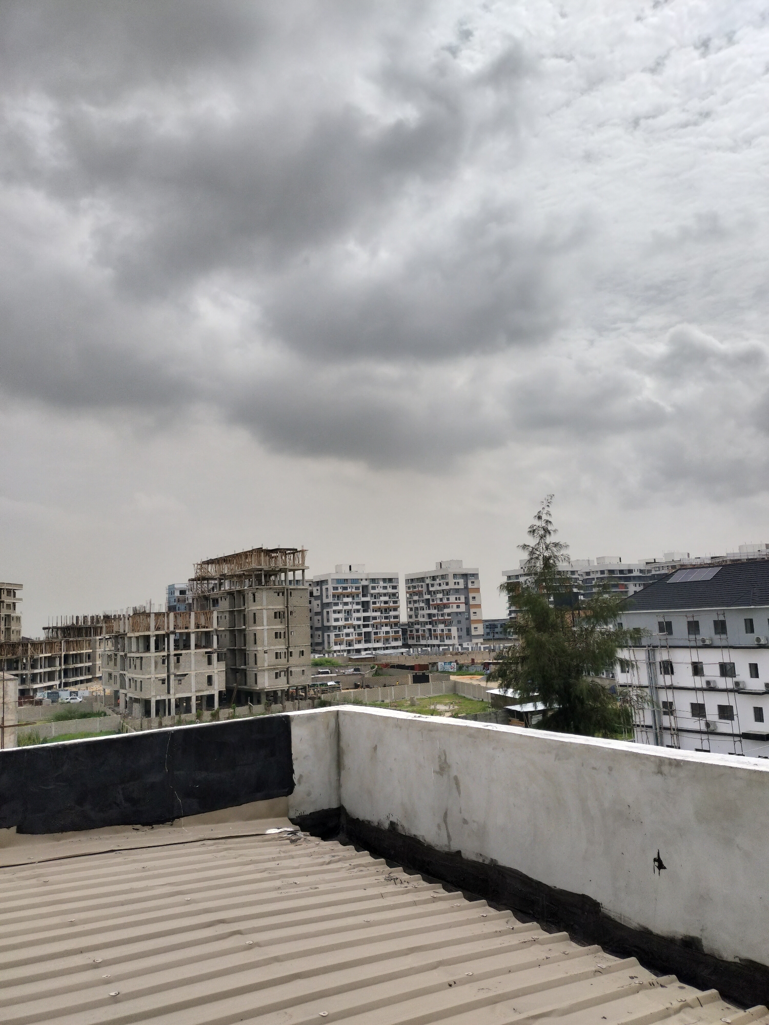 The image depicts a view from a rooftop in Moba, Nigeria. In the foreground, a corrugated metal roof and a low concrete retaining wall are visible. Behind the wall, a cityscape unfolds under a heavily overcast sky. Several multi-story buildings are under construction, their concrete structures exposed. Other completed buildings are visible, some with white facades and visible windows. A single tree stands out amidst the buildings. The overall impression is one of urban development in progress under a cloudy sky.