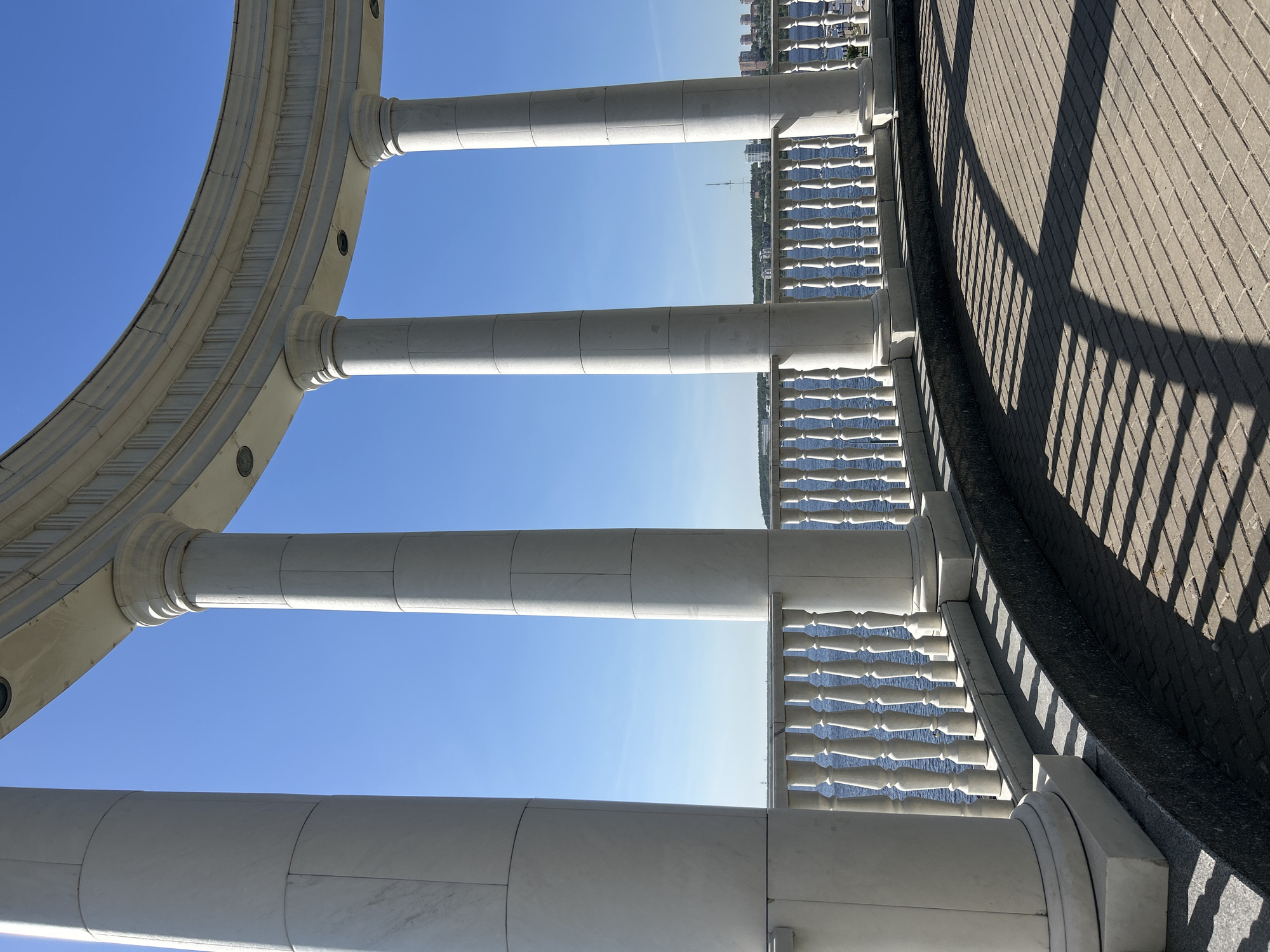 The image shows a close-up, low-angle view of a colonnade in Izhevsk, Russia. The colonnade consists of white, cylindrical columns capped by a curved, ornate architrave. The columns frame a view of the Kama River, with a distant shoreline featuring buildings and foliage. A white balustrade runs along the edge of the colonnade, partially obstructing the view of the river. The foreground includes a section of paved walkway, with shadows cast by the balustrade and columns. The sky is clear and blue.