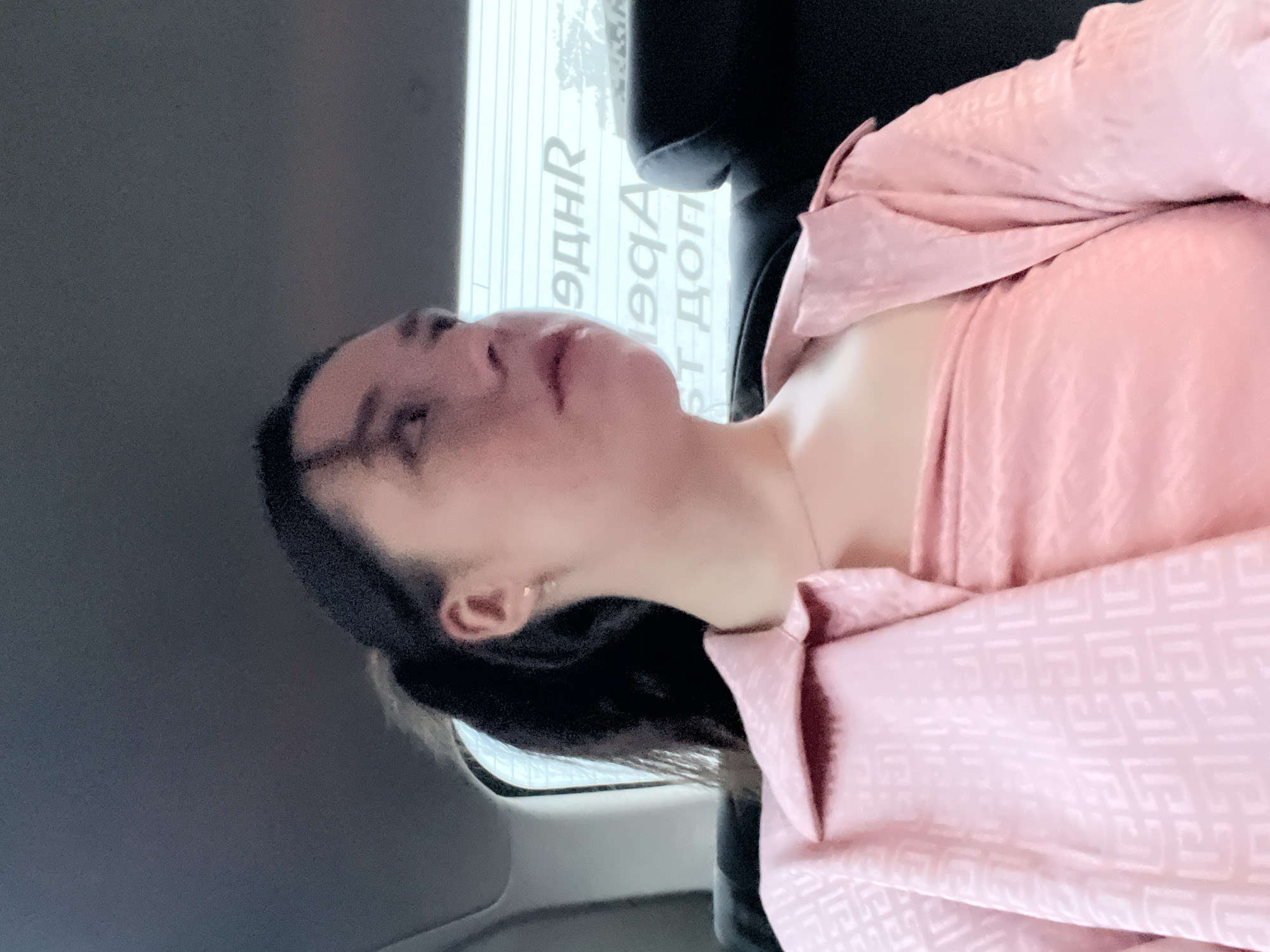 A woman with dark hair is wearing a pink patterned blouse and is positioned inside what appears to be a car. The background shows a window with text, possibly signs in Cyrillic script, suggesting the location is likely Moscow, Russia.
