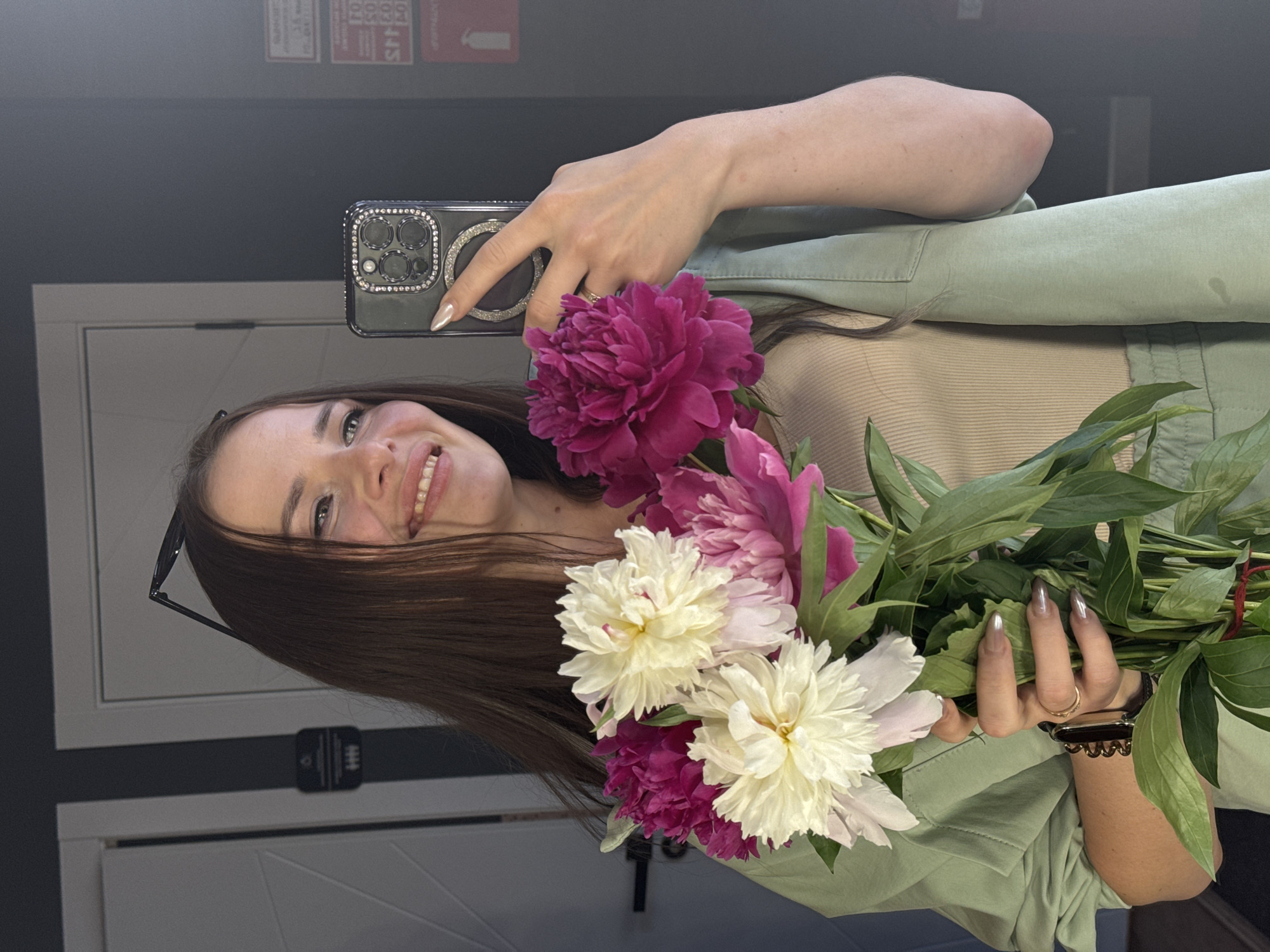 A young woman with long brown hair is taking a mirror selfie in what appears to be a domestic or personal space. She is holding a bouquet of peonies, with white and pink flowers prominently displayed. She is wearing a light green jacket over a beige top. Her phone, adorned with rhinestones, is visible in her right hand as she takes the picture. She is smiling and looking at her reflection. The background shows doors and a dark wall with some signage. The location is identified as Chaikovsky, Russia.