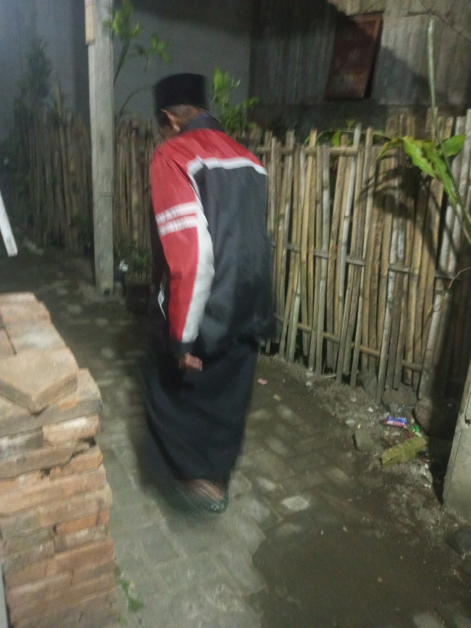 The image depicts a person wearing a black peci and a red and black jacket over a long black garment, possibly a sarong, walking away from the camera in a dimly lit outdoor environment. The person is walking on a wet paved surface, and to the right, a bamboo fence is visible. To the left, a stack of bricks is present. In the background, a rough wall and some greenery are discernible. The scene appears to be an alleyway or a pathway in a residential area. The lighting suggests it is nighttime. The location is identified as Andongsili, Indonesia.