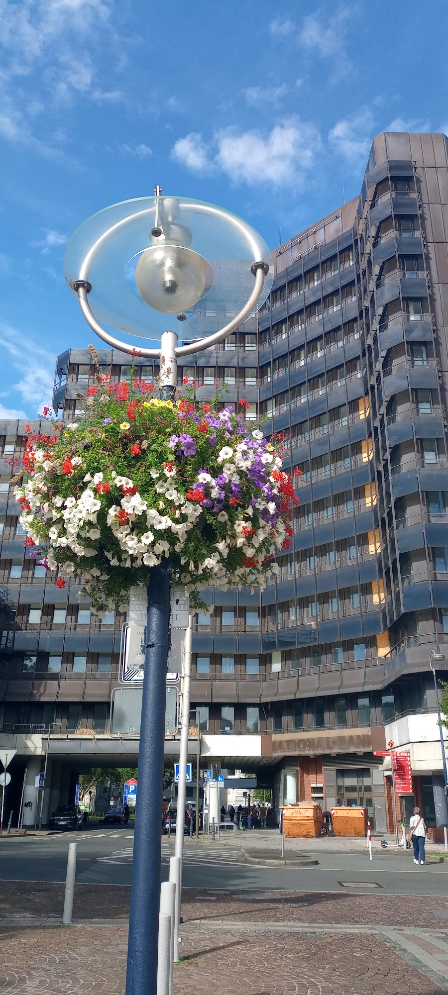 The image shows a street scene in Dortmund, Germany. In the foreground, a tall, dark blue lamppost is adorned with a large, overflowing basket of colorful flowers, including white, red, and purple petunias. Above the flowers, the lamppost features a circular, clear glass shade with a metallic frame, and a bulb is visible within. Behind the lamppost, a large, modern building with a dark facade and numerous windows dominates the background. The building appears to be an office building or bank. A street with crosswalks and a few vehicles is visible in the middle ground. Several pedestrians are walking on the sidewalk in front of the building. Two large orange dumpsters are placed on the sidewalk. The sky is blue with scattered white clouds. The location is identifiable as the area in front of the former "National-Bank" building, which is part of the Reinoldiplatz in Dortmund, Germany.