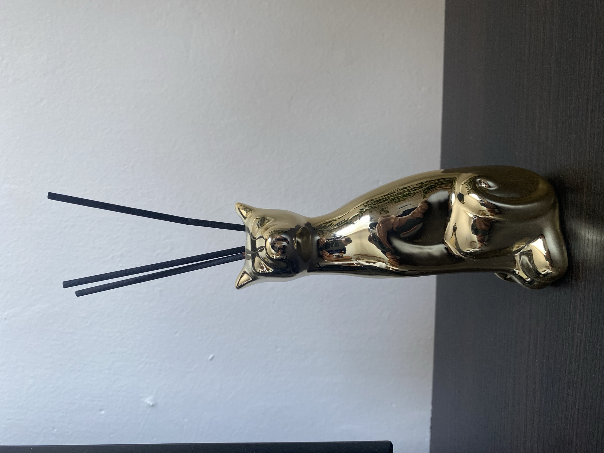 The image shows a golden cat statue with two black incense sticks inserted through its body. The cat is in a lying down pose and is highly reflective, showing glimpses of what appears to be a person taking the photo. The statue is positioned against a wall that is split between a white textured surface on the left and a dark wood-grained surface on the right. The lighting suggests an indoor setting, possibly a room. There is no visible text in the image, and no specific time of day or weather conditions are discernible. The overall scene is static, focusing on the decorative object and its placement.