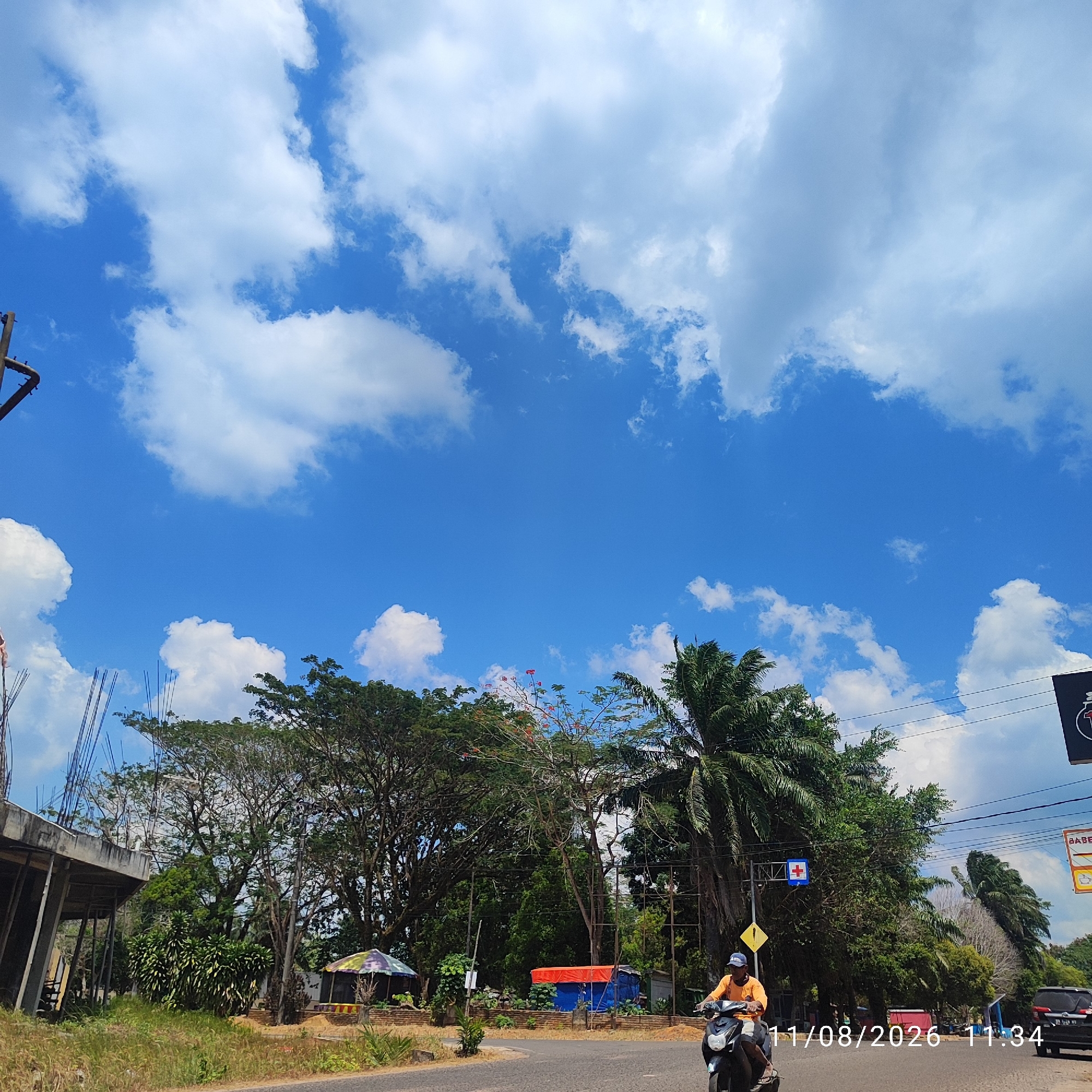 The image depicts a street scene in Buding, Indonesia, under a bright blue sky with scattered white clouds. In the foreground, a person is riding a motorcycle on a paved road. The rider, wearing an orange shirt and a dark cap, is facing to the left. To the right of the road, a black SUV is visible.
The middle ground of the image is characterized by lush green trees and vegetation, with some buildings and construction in the background. On the left, an unfinished concrete structure with exposed rebar suggests ongoing construction. Further back, a colorful, umbrella-like structure stands out, possibly a stall or shelter. A few utility poles and wires are also present. A blue sign with a red cross is mounted on a pole, indicating a medical facility or emergency services.
The overall environment suggests a tropical or sub-tropical climate, with abundant plant life and a clear sky. The presence of the construction site and the traffic indicates a lived-in urban or semi-urban area. A timestamp in the lower right corner reads "11/08/2026 11:34".