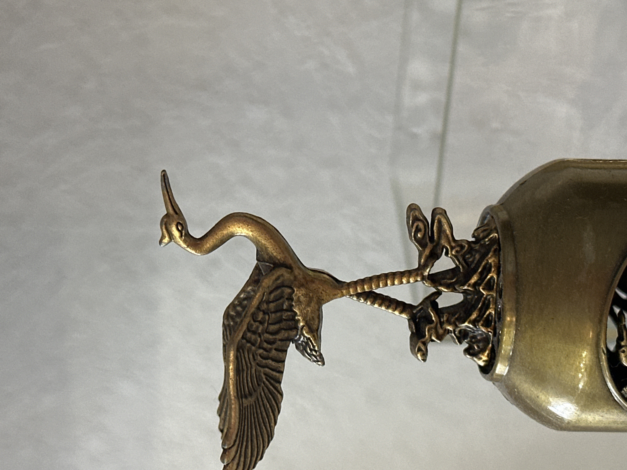 A close-up, eye-level, slightly tilted shot captures a decorative brass figurine of a crane standing on what appears to be stylized rocks or tree roots atop a metallic, bulbous object. The crane is golden-bronze with intricate detailing on its wings, body, and long legs. Its head is slightly raised, and its beak is pointed forward. The background is a plain, textured light gray surface, possibly a wall or fabric. The object below the crane has a rounded, metallic surface with a visible circular cutout revealing a dark interior with some ornamentation. The lighting is soft, creating subtle highlights and shadows on the metallic surfaces. The image is sharp and focuses on the figurine and the upper portion of the object it rests on. There is no visible information in the image that would indicate its location in Nevinnomyssk, Russia.