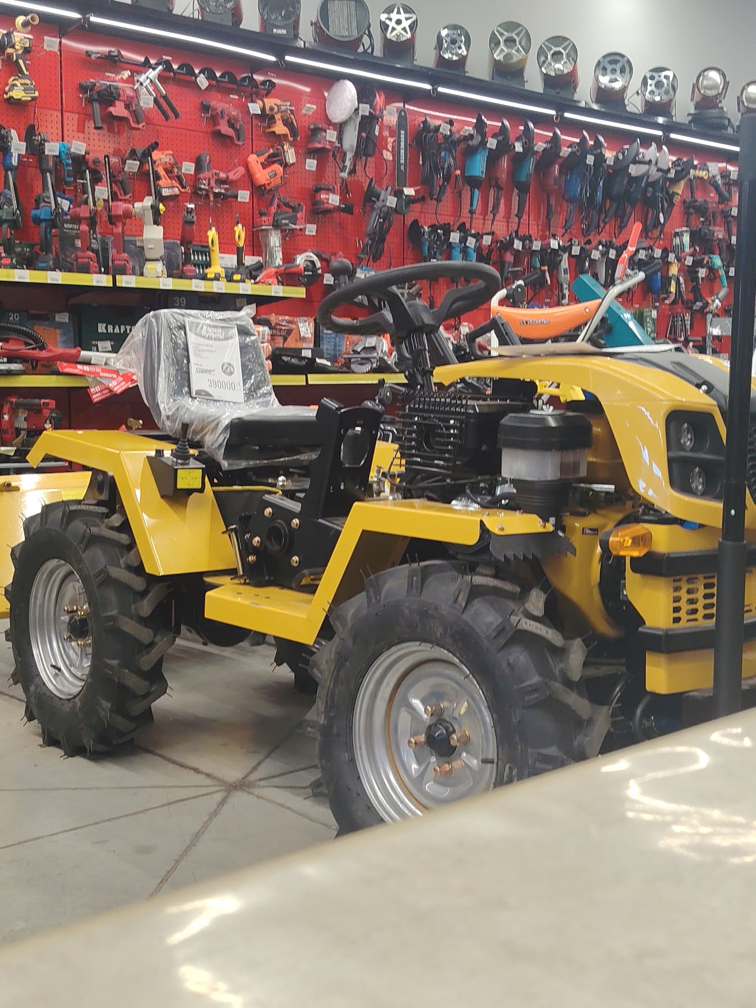 The image depicts a yellow tractor, possibly a walk-behind model, parked indoors. The tractor has large, knobby tires, a black seat covered in plastic, and a visible engine. The background reveals a wall lined with various tools, including drills, saws, and other power tools, suggesting the location is a retail store or hardware store. The lighting is consistent with indoor retail environments. There are no people visible in the frame, nor any indication of the city or specific location beyond being an indoor retail setting.