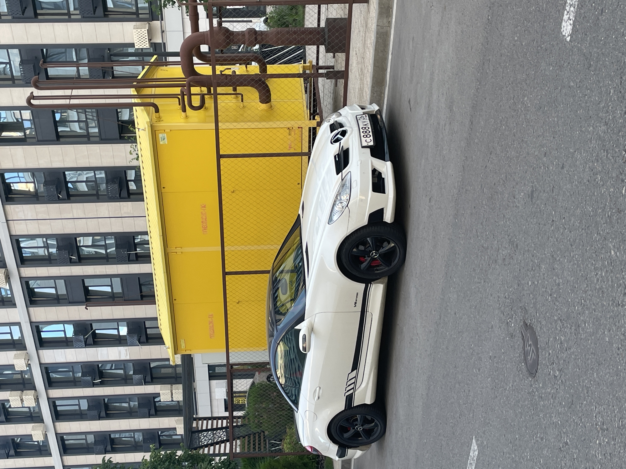 In Stavropol, Russia, a white convertible Mercedes-Benz SLK-Class is parked on an asphalt road in front of a modern building with cream-colored facade panels and dark-framed windows. To the right of the car is a bright yellow industrial enclosure with a metal fence in front of it, behind which are large diameter rusty pipes. The car has black wheels with red brake calipers and black racing stripes along its side. A license plate on the front of the car reads "C 888 KY 26". A circular metal manhole cover with an embossed symbol is visible on the road in the foreground.