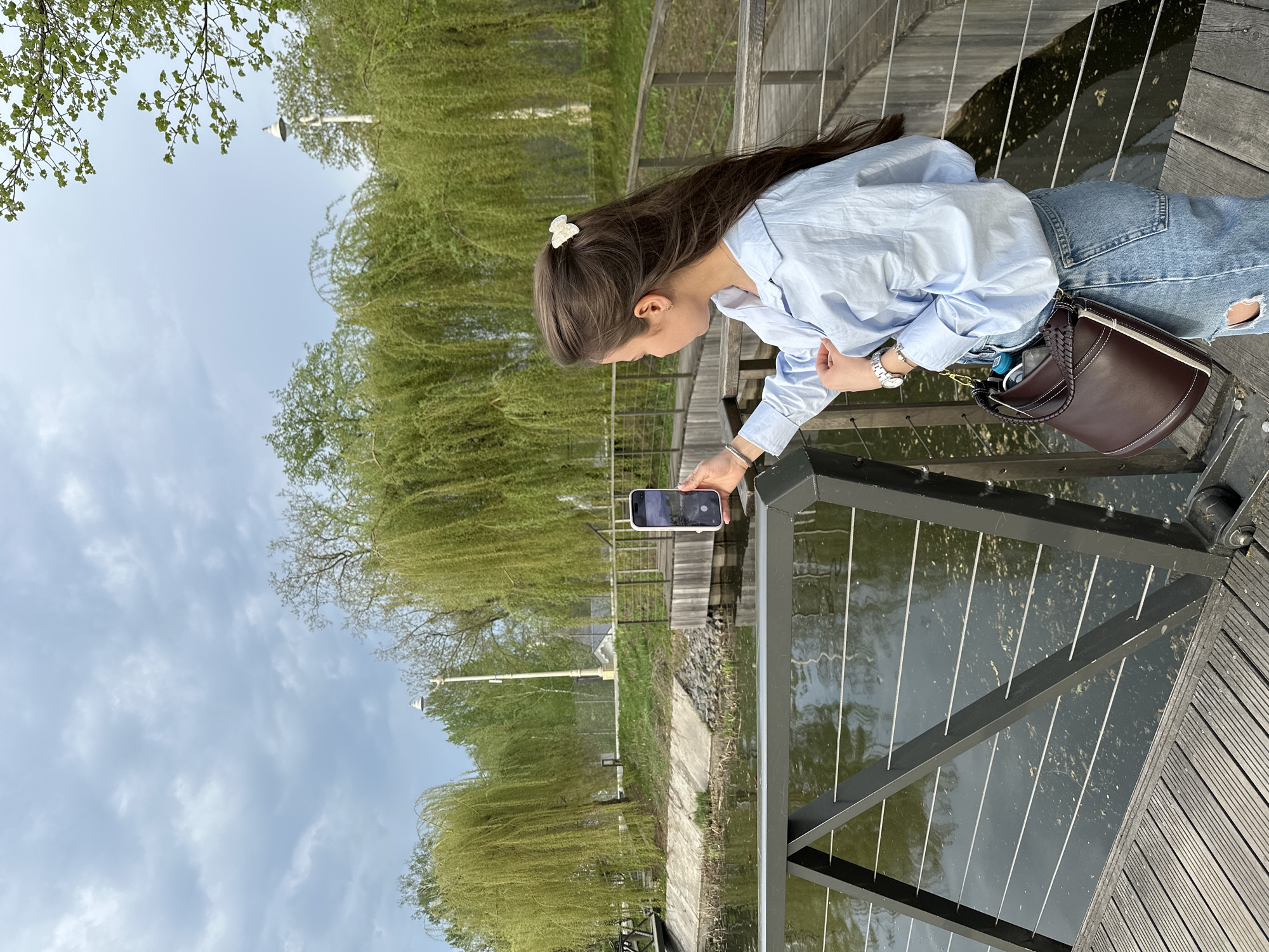 A young woman stands on a wooden boardwalk alongside a body of water, potentially a pond or river, in an outdoor setting. She is wearing a light blue shirt, blue jeans, and a wristwatch. Her dark hair is adorned with a white butterfly hair clip. She is holding a smartphone, apparently taking a picture of the water. A dark brown handbag is hanging off her shoulder. The background features trees, some with weeping branches, and a cloudy sky. A lamppost is visible in the distance. The location is possibly a park or recreational area in Moscow, Russia.
