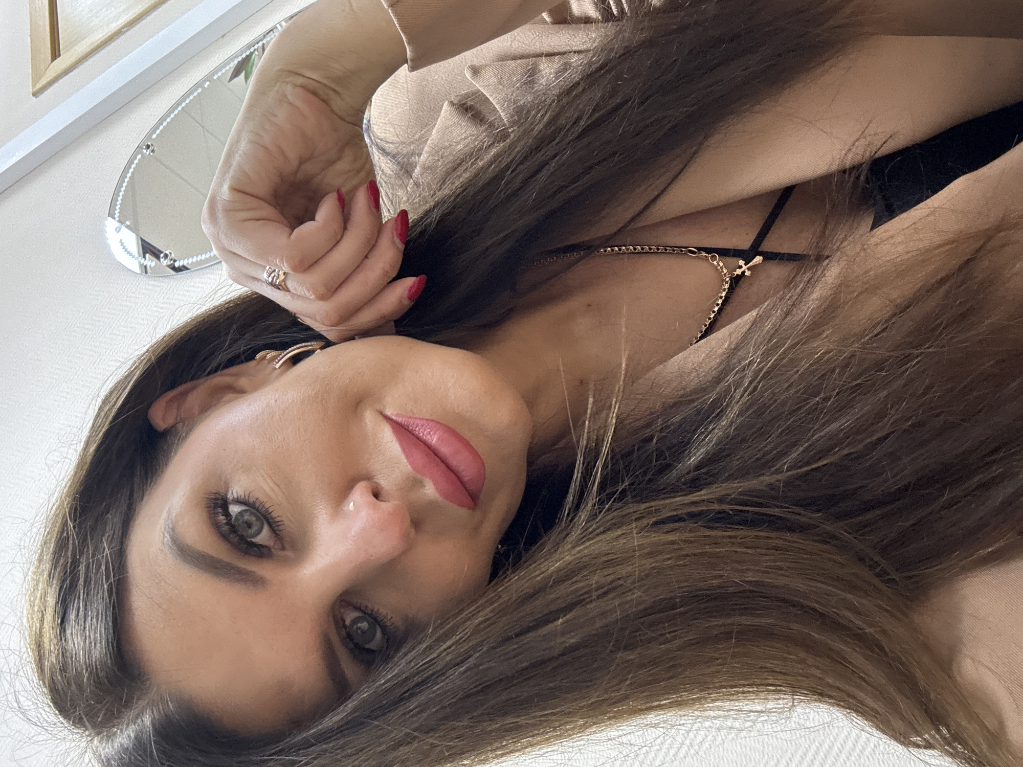 A woman with long, brown hair and prominent makeup poses for a selfie. She is wearing a light brown blazer over a black top, and a gold chain with a cross pendant. Her left hand is resting on her cheek, and she is wearing a ring on her index finger and hoop earrings. A small nose stud is visible. The background appears to be an indoor setting with a textured wall and a decorative mirror reflecting part of the room. The image does not contain any identifiable landmarks or elements that would suggest a specific location within the city of Chaikovsky, Russia.