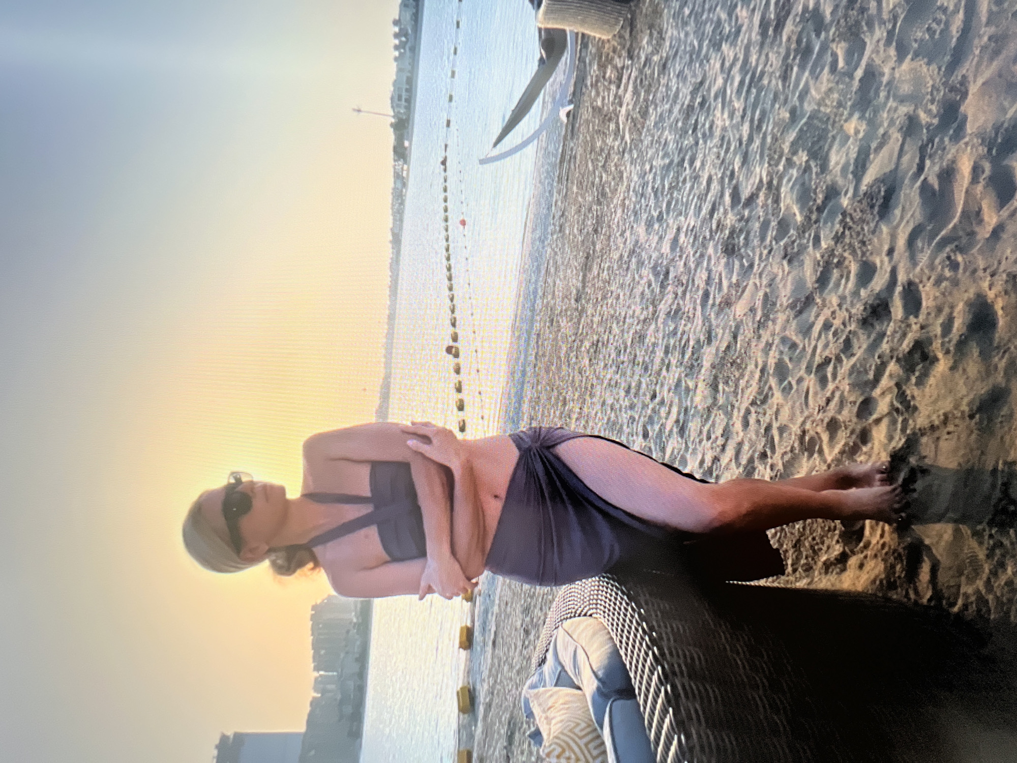 A medium shot captures a woman standing on a sandy beach at sunset. She is wearing a grey bikini top and a matching sarong tied at her hip, with a high slit revealing her left leg. Her arms are crossed, and she gazes to her right, away from the camera. She has blonde hair pulled back and is wearing dark sunglasses. To her left, a portion of a dark wicker couch with cushions is visible. The background shows the calm, light blue ocean stretching to the horizon, with a city skyline faintly visible in the distance on the left. The sky is a warm gradient of yellow and blue, indicative of sunset. A buoy line stretches across the water.