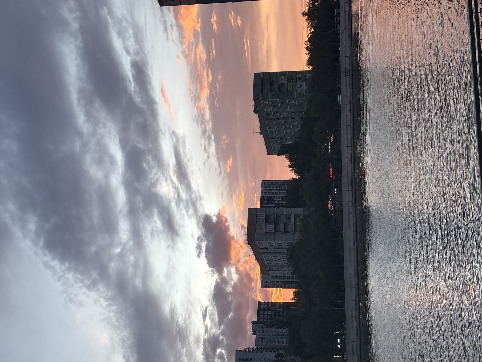 A vertical photograph captures a sunset over a cityscape with a body of water. The sky on the left is filled with dramatic, gray clouds, while the right side features a dramatic sunset with streaks of orange and pink. Silhouetted against the vibrant sky are several tall, residential buildings, their windows visible. Trees line the waterfront between the buildings and the water. The water, on the right side of the image, reflects the colors of the sunset, creating a textured, shimmering surface. The scene suggests an urban environment at dusk, likely during a period of fair weather with interesting cloud formations. No people are visible. There is no discernible text in the image.