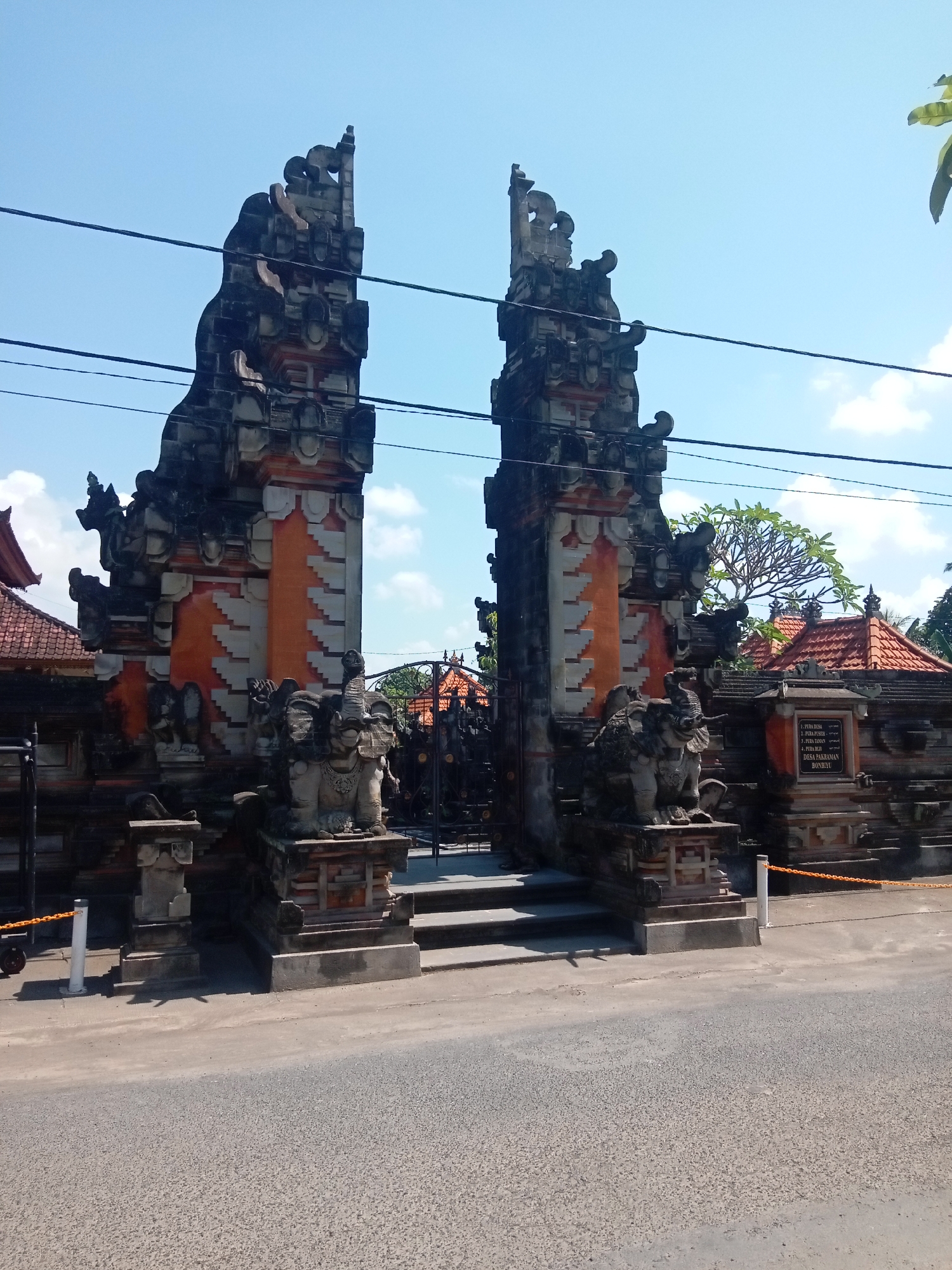 The image shows an ornate, intricately carved stone gateway in an Asian architectural style, likely Balinese, set against a bright blue sky with scattered clouds. The gateway is flanked by two tall, multi-tiered towers, each topped with decorative elements resembling curling motifs. Between the towers, a dark metal gate is visible, with steps leading up to it. On either side of the entrance, at the base of the towers, are stone sculptures of elephants, facing outward. The gateway is constructed with a combination of dark and light stone, with sections of vibrant orange and lighter patterned stone cladding on the towers.
To the left and right of the central gateway, a wall with traditional red-tiled roofs is visible, suggesting this is the entrance to a complex, possibly a temple or cultural site. There are also decorative elements on the wall, including what appears to be a carved plaque with text, though the text is not legible in the image. The gateway is situated next to a paved road, with some yellow and orange safety chains and posts marking the edge of the property. Power lines are visible crossing the sky above the gateway. The overall impression is of a grand and culturally significant entrance.