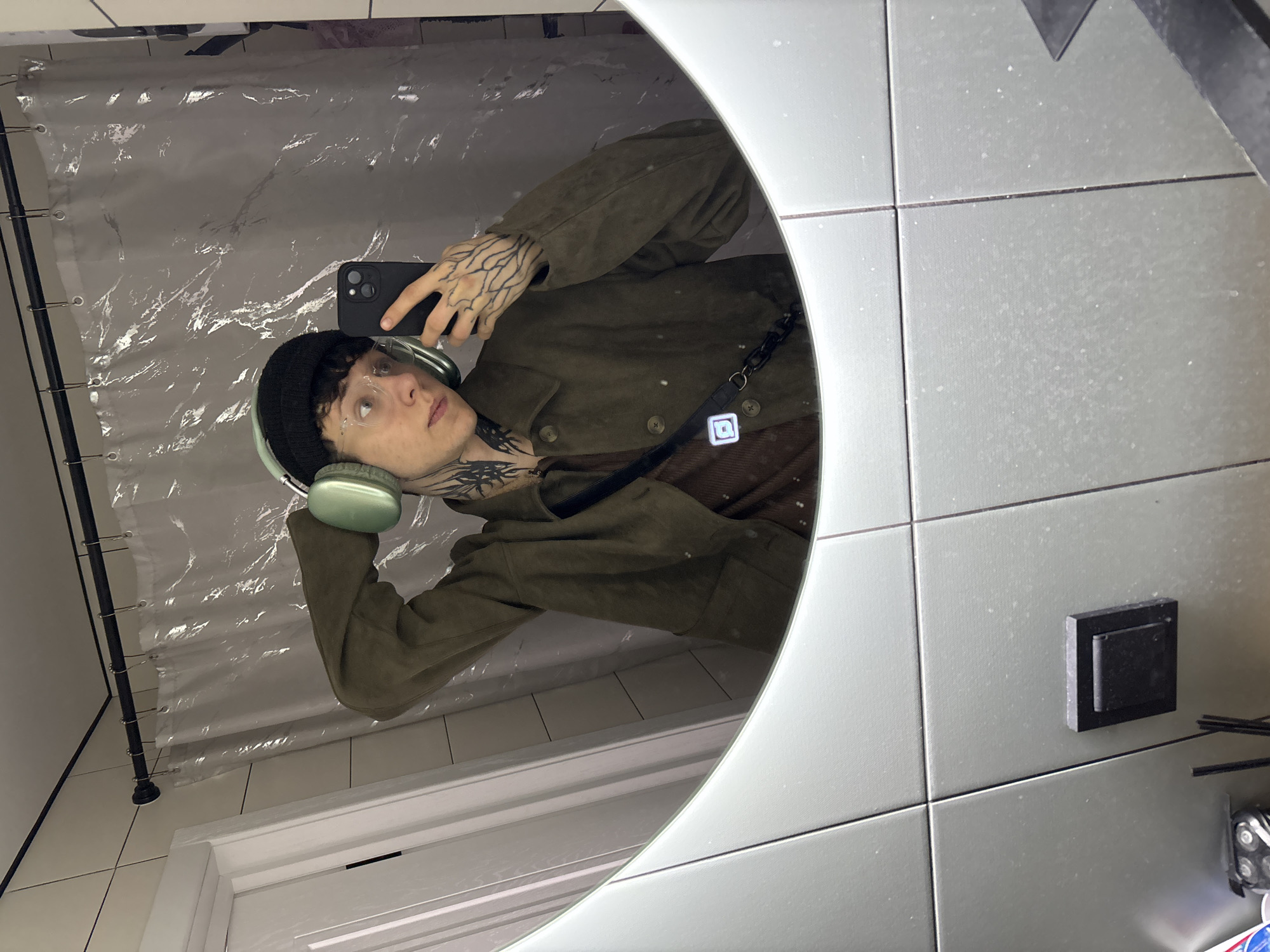 The scene is located in a bathroom in Saratov, Russia. An individual is captured in a mirror selfie, seen from the chest up. The individual wears a black beanie, clear-framed glasses, and light green over-ear headphones. They are dressed in a dark olive-green collared jacket over a brown undershirt. Dark, intricate tattoos are visible on the left side of the neck and cover the entire visible portion of the left hand, resembling root or vein patterns. A black strap, possibly from a bag, is visible across the individual's chest. The left hand holds a black smartphone with a triple-lens camera array, pointed towards the mirror to capture the image. The right hand is raised towards the right side of the head, appearing to adjust the headphones. The background, reflected in the mirror, includes a translucent, crinkled plastic shower curtain hanging from a black rod with metal rings. Behind the curtain, white rectangular tiles with dark grout lines are visible. A white doorframe is partially visible to the left. The lower perimeter of the mirror is bordered by grey tiled walls with dark grout lines, showing minor speckling. A black square electrical switch or outlet is mounted on the grey tiles below the mirror. Several narrow, dark cylindrical objects are visible on an unseen surface beneath the mirror.