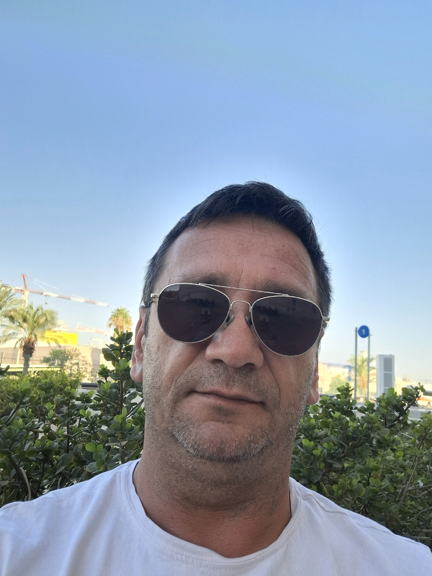 A man wearing aviator sunglasses and a white t-shirt is photographed outdoors, against a clear blue sky. He has short, dark hair and stubble. In the background, green foliage is visible, along with palm trees and construction cranes. A building with yellow and white elements can be seen behind the trees, and a blue and white sign is also present. The setting appears to be an urban environment in Kiryat Gat, Israel, likely during daylight.