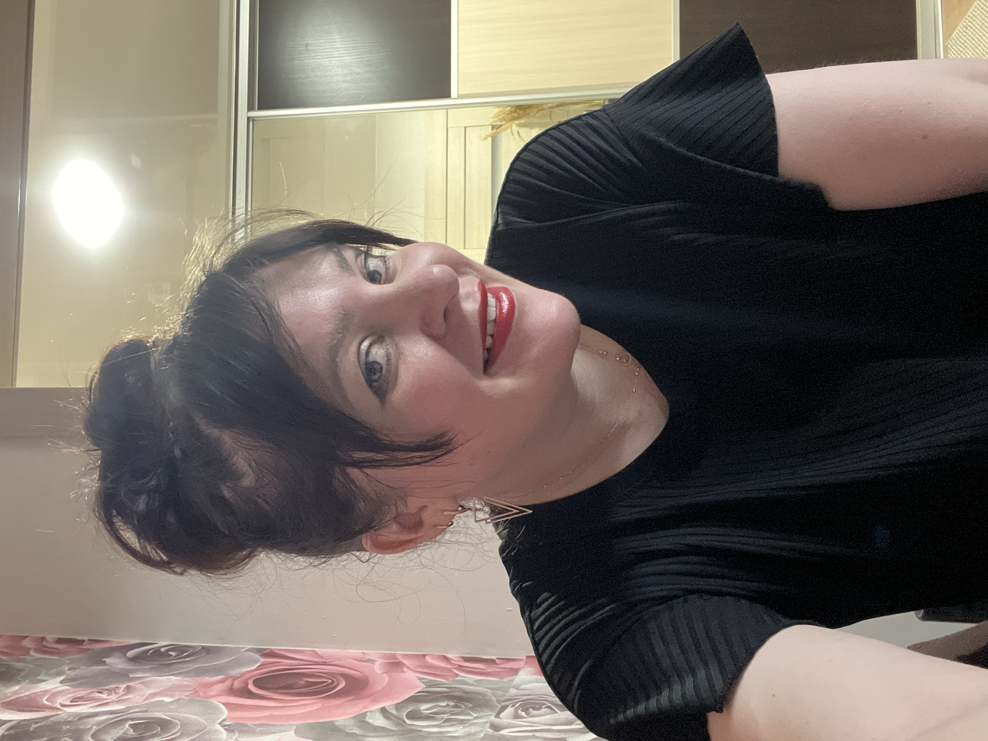 A single adult female is depicted in a close-up, head-and-shoulders portrait shot within an indoor residential setting in Noviy, Russia. The subject has dark hair styled in a high bun with loose strands framing her face, light-colored eyes, and is wearing red lipstick while smiling directly at the camera. She is wearing a black, ribbed short-sleeved top, small triangle-shaped earrings, and a delicate necklace. The background to the left features a wall covered with wallpaper displaying large floral patterns in shades of pink, grey, and white. To the right, a multi-paneled sliding door wardrobe or closet is visible, composed of light and dark wood-grain sections, with a bright, rectangular overhead light fixture reflected in its upper surface. A small bundle of dried natural material, resembling grass or reeds, stands against the wardrobe in the lower right background.