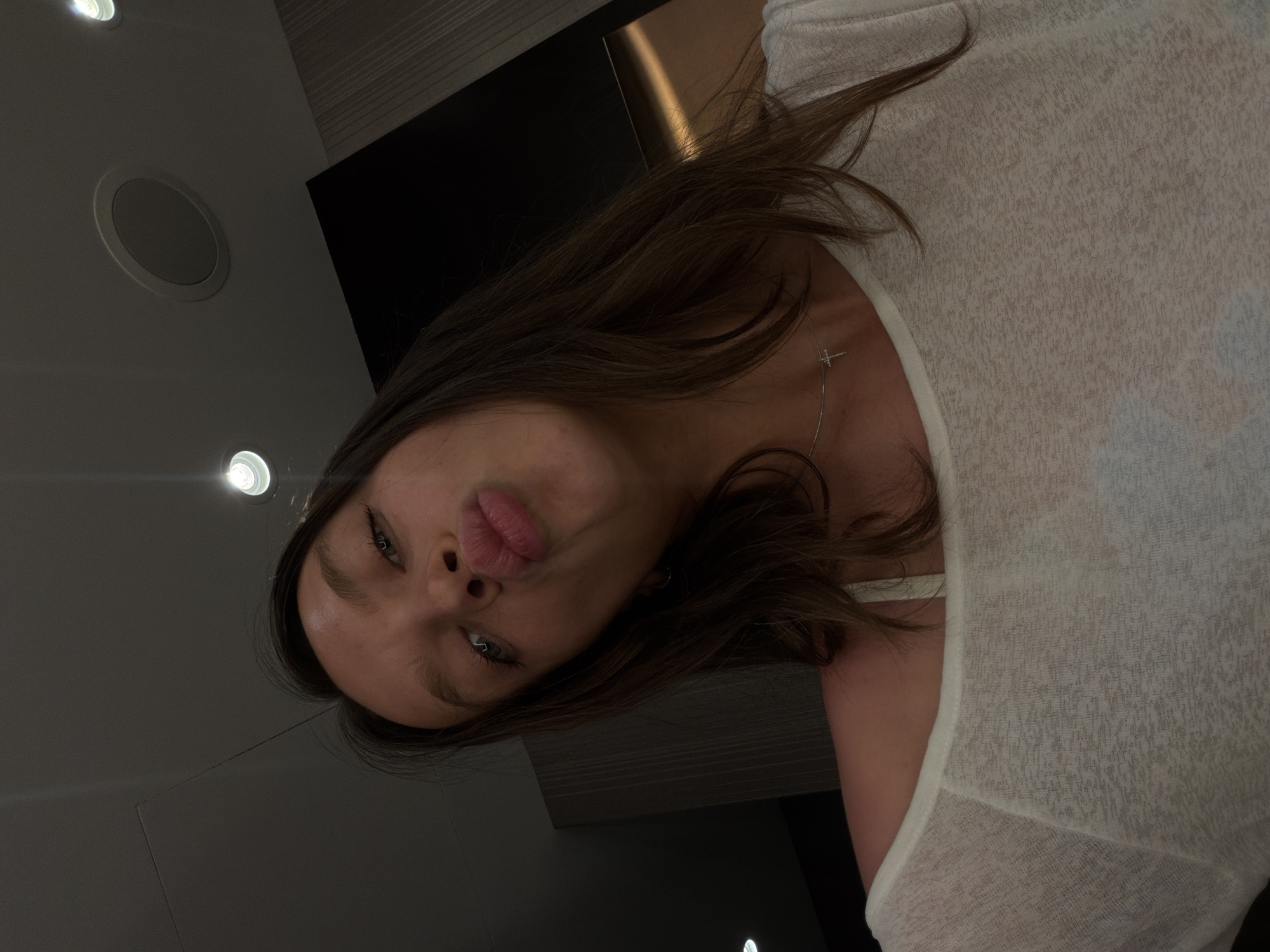The image depicts a low-angle selfie of a young adult female, approximately in her late teens or early twenties, with long dark brown hair and light skin. Her blue-grey eyes are directed upwards, and her lips are pursed into a "kissing" expression. She is wearing a sheer, loose-fitting, light-colored long-sleeved top that reveals a light-colored bra strap on her right shoulder. A thin, silver-colored necklace with a small cross pendant is visible around her neck. The setting is an indoor room in Budva, Montenegro, characterized by a dark, possibly paneled wall on the left and a lighter, textured or striped wall on the right. The ceiling features several recessed circular light fixtures, some illuminated, and a central circular vent or speaker cover. The overall lighting is subdued, with artificial light sources from the ceiling providing illumination.