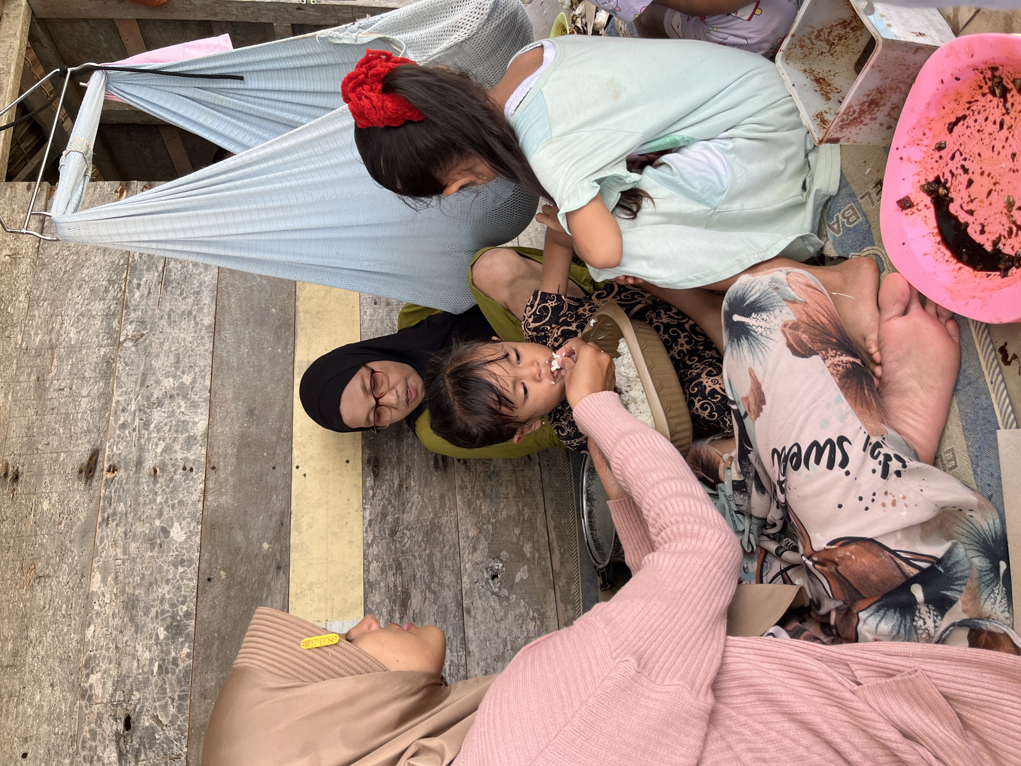 In Dayahtuha, Indonesia, a group of people are gathered on the ground in what appears to be an outdoor setting. The background consists of weathered wooden planks. A woman in a light pink knitted cardigan and a beige hijab is feeding a young child, who is wearing a dark patterned outfit, from a bowl of white rice. Beside them, another woman in a black hijab and glasses is seated. To the right, a young girl with a red hair accessory is also present. In the background, a light blue hammock is suspended. Various containers and food items are scattered around the group.
