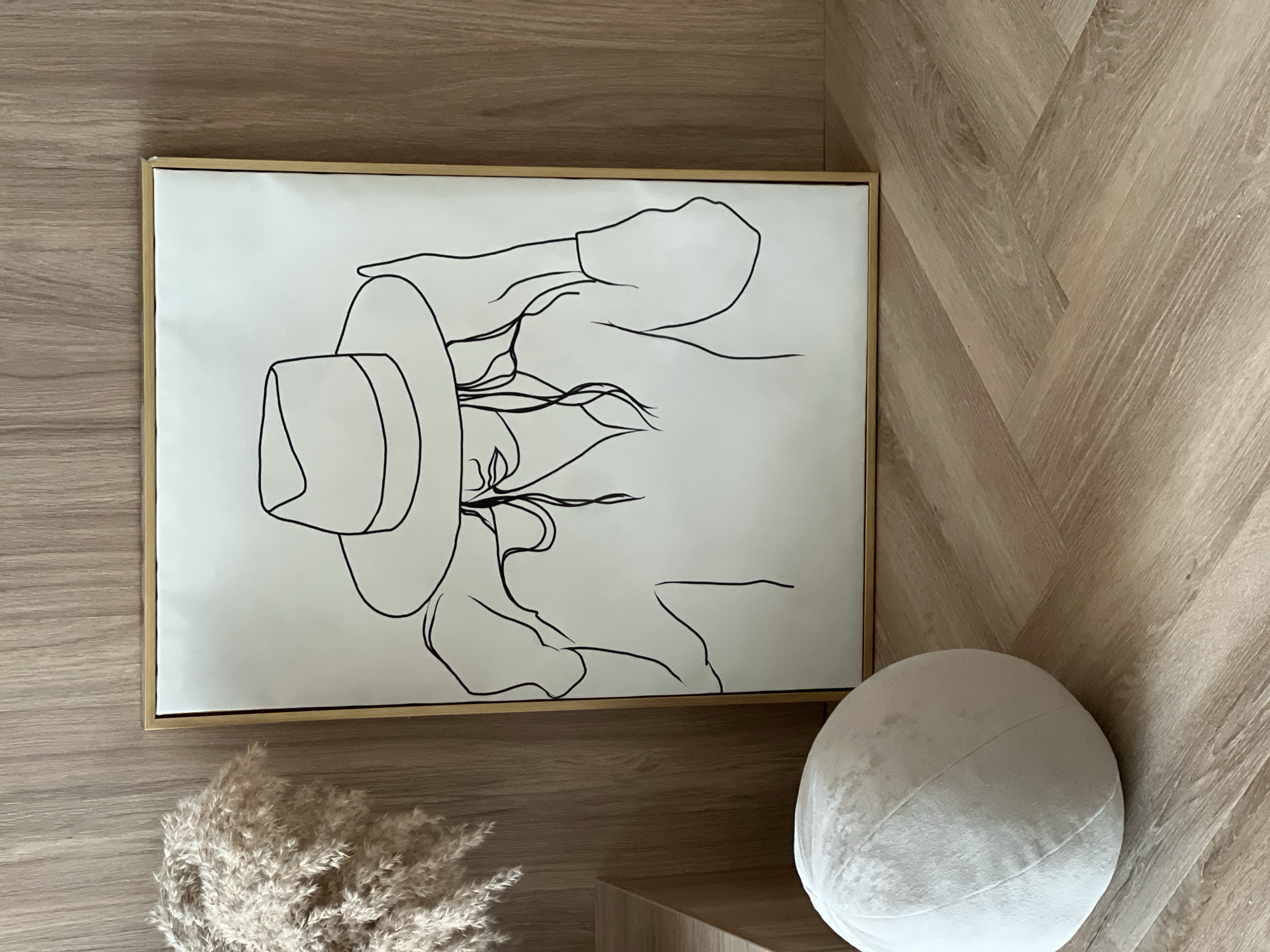 The image displays an indoor arrangement featuring decorative objects. A large, framed piece of abstract line art is positioned centrally, leaning against a wall or resting on a surface. The artwork, a minimalist black line drawing on a white background, depicts the upper body of a person, likely a woman, wearing a wide-brimmed hat, with hair flowing out from under it. The frame around the art is thin and gold-colored.
To the lower left of the frame, a bunch of light-colored, fluffy dried pampas grass or a similar botanical element is visible. To the lower right, a round, white, textured cushion or decorative sphere is placed. Both the pampas grass and the sphere are situated on a wooden surface, possibly a low shelf or cabinet. The entire setup rests on a light-toned wooden floor with a distinct herringbone pattern.
The scene is static, depicting no specific activity or event. The lighting appears soft and ambient, characteristic of an indoor environment. There are no visible cues indicating the time of day or specific outdoor weather. No text is visible anywhere in the image.
