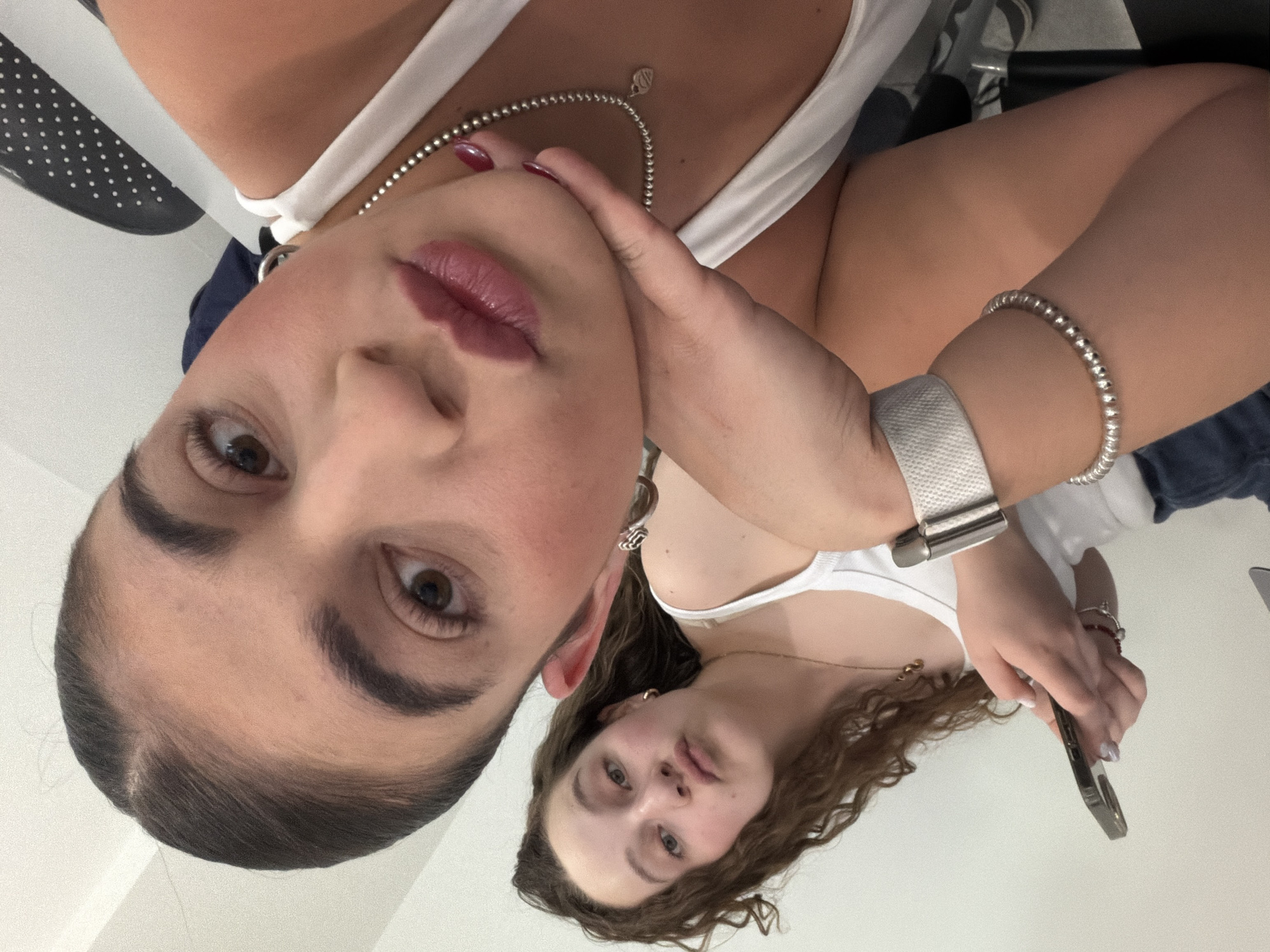 This image captures two young women in what appears to be a mirror selfie, or a photograph taken from an unusual, low angle, resulting in a 90-degree clockwise rotation of the scene. The foreground features a woman with dark, slicked-back hair and brown eyes, looking directly into the camera. She has prominent eyebrows and full lips with a dark pink or plum-colored gloss. She wears a white tank top, a silver beaded necklace with a small pendant, and a large silver hoop earring in her left ear. Her right hand, adorned with red nail polish, is gently placed against her chin. On her right wrist, a silver beaded bracelet is visible, and on her left wrist, a white watch strap with a silver clasp can be seen.
In the background, slightly out of focus, is another woman with long, curly brown hair and lighter eyes. She is also wearing a white tank top and a delicate gold necklace. She holds a dark-colored smartphone in her right hand. The setting is indoors, characterized by bright, even lighting and a largely white background, possibly a wall or reflective surface. A small portion of a dark object with white polka dots is visible in the upper left corner. The subjects' expressions are neutral. No visible text is present in the image. The location context for this media file is San Bernardino Tlaxcalancingo, Mexico.