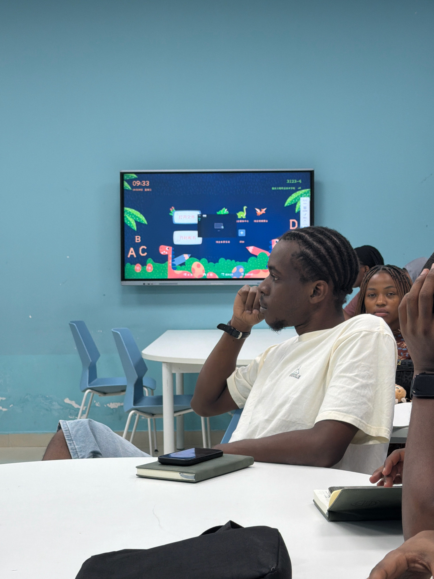 In a brightly lit room with pale blue walls, a group of young adults are seated around a white circular table, likely in a classroom or learning environment in Jiangjin, China. A large digital display mounted on the wall shows educational content, featuring cartoon dinosaurs, letters, and Chinese characters. In the foreground, a young man with braided hair, wearing a light-colored t-shirt, is looking to the side, his hand near his chin. He is seated at the table, which has a notebook and a smartphone on it. To his right, another young woman with braided hair is looking forward, appearing somewhat thoughtful. The room also contains several blue plastic chairs and what appears to be a partition separating different sections of the space. The overall atmosphere suggests a contemporary educational setting.