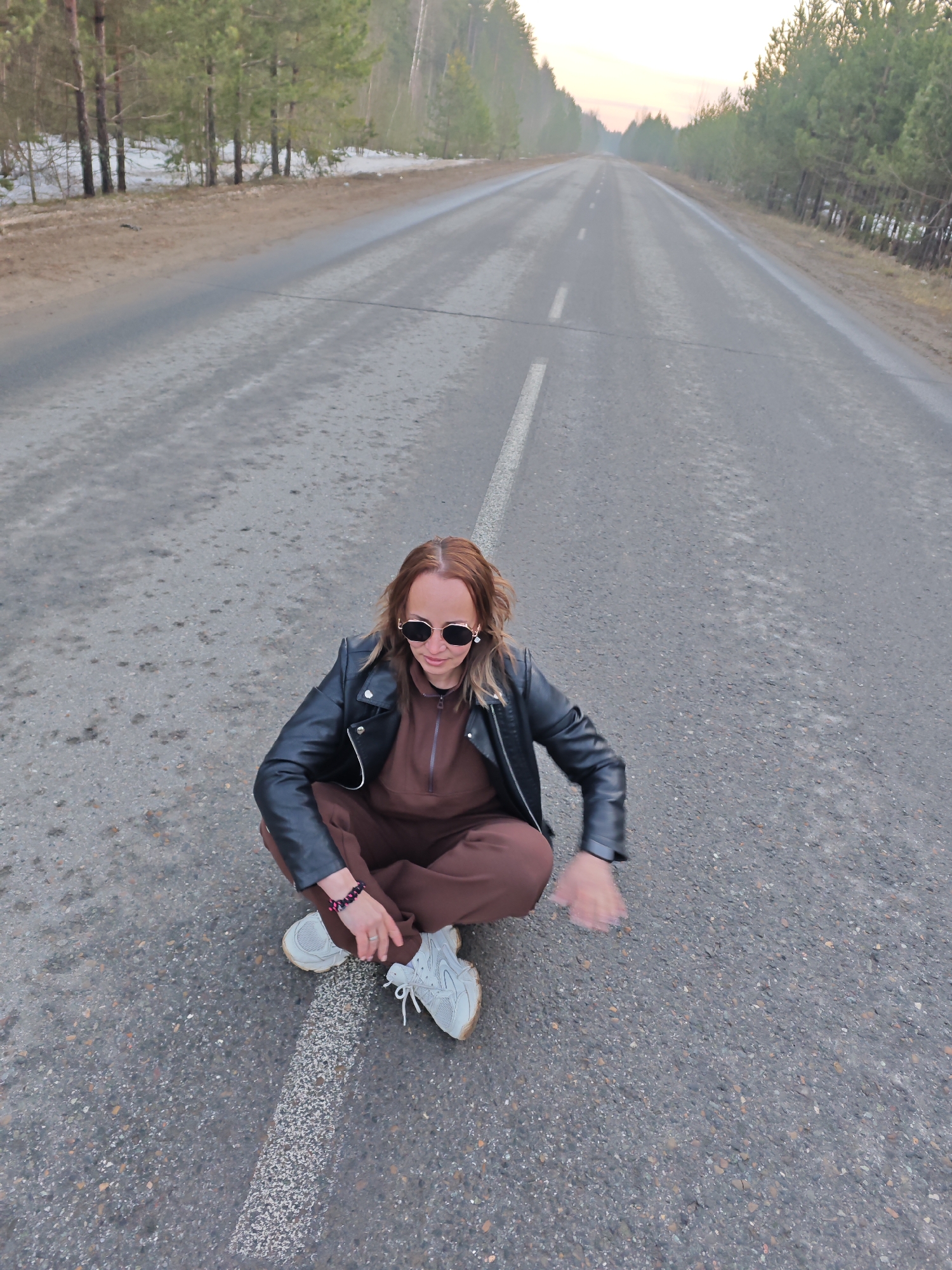 A woman with light brown hair and sunglasses sits cross-legged on a paved road marked with a white line. She wears a brown jumpsuit and a black leather jacket, along with white sneakers and a bracelet on her left wrist. The road stretches into the distance, flanked by trees on both sides, with lingering snow visible along the edges. The sky is a pale, early evening hue. The location is in the city of Chaikovsky, Russia.