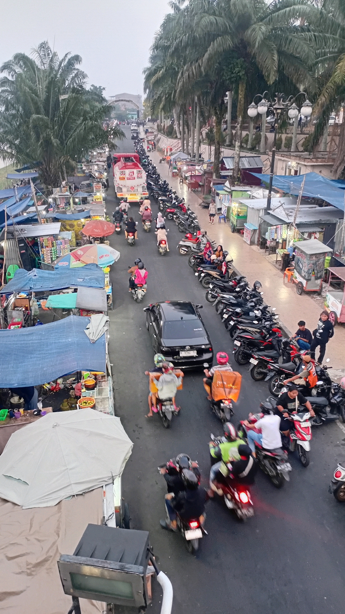 The image depicts a busy street scene in Jambi, Indonesia, likely near the Batanghari River given the presence of palm trees and market stalls often found near waterways in tropical Indonesian cities. The perspective is from an elevated vantage point, looking down on a two-lane road filled with a variety of vehicles, primarily motorcycles. On the left side of the road, a market or roadside stalls are set up with blue and gray tarps covering various goods, including food items and drinks. Some vendors are visible behind their stalls.
The road is congested with traffic. A red fuel tanker truck is prominent in the upper-left portion of the image. Numerous motorcycles are in motion, some carrying single riders and others with passengers. Several parked motorcycles line the right side of the road, indicating a popular area or destination. A black sedan is navigating the traffic.
People are interacting within the scene: some are riding motorcycles, others are walking near the stalls, and a group of men are seated on motorcycles on the right side of the road. The environment appears to be an urban setting with a row of tall palm trees lining the right side of the street, suggesting a tropical climate. The sky is overcast, casting a diffused light over the scene. The overall impression is one of a vibrant, active street market and transportation hub in Jambi.