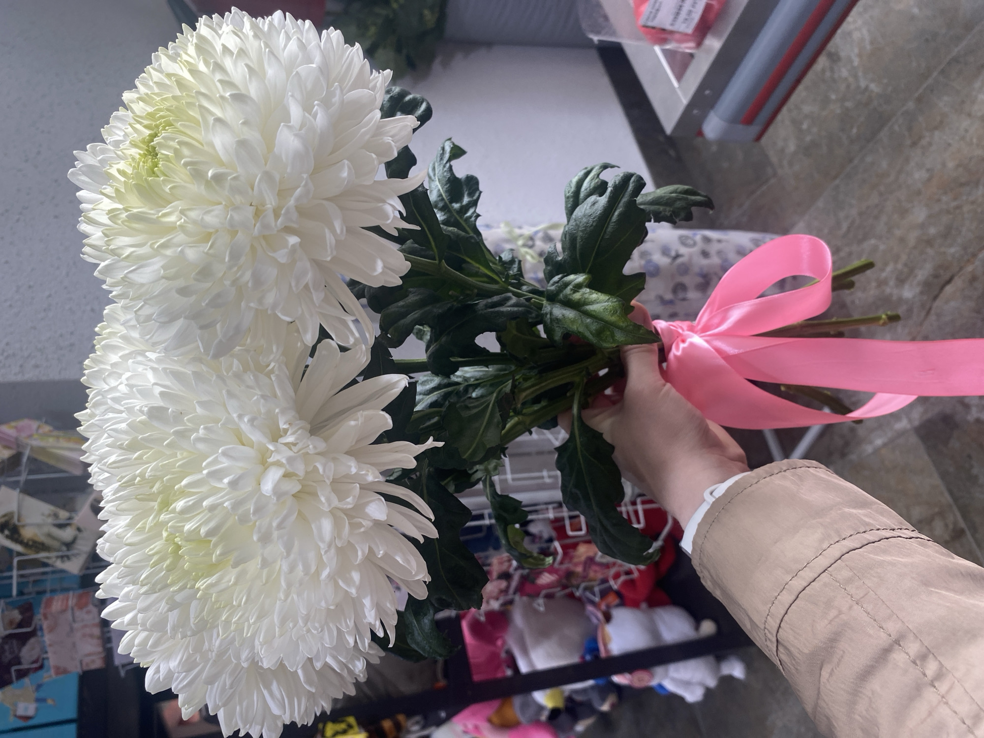 A hand holds a bouquet of white chrysanthemums tied with a pink ribbon. The background is blurry, but hints at a store environment, possibly in Yekaterinburg, Russia, indicated by retail shelving and merchandise.
