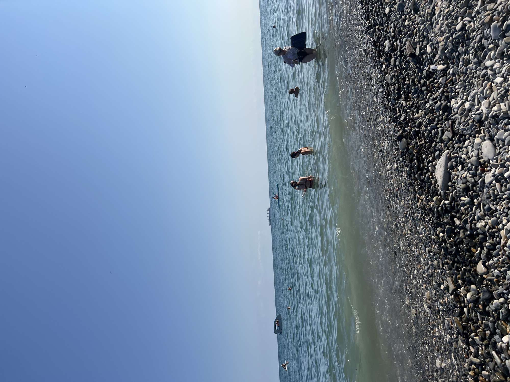The image depicts a clear, sunny day at a pebble beach on the Black Sea coast, likely in Tuapse, Russia. The foreground is dominated by a dense collection of smooth, grey, black, and white pebbles. Gentle waves are lapping at the shore, creating a foamy edge.
In the water, several people are visible. To the right, a woman wearing a hat and a dark swimsuit is standing in the shallow water, holding a dark rectangular object, possibly a boogie board or a kickboard. Further out, two young women are wading in the water, one with her back to the camera, wearing a dark swimsuit, and another facing the camera, also in a dark swimsuit. In the mid-ground, a person is partially submerged in the water, appearing to be swimming.
In the background, the calm, blue sea stretches to the horizon under a bright, clear blue sky. A few distant objects are visible on the water, including a small boat or inflatable watercraft, a larger ship on the horizon, and possibly another person on a paddleboard. The overall scene suggests a recreational activity at a popular beach location.