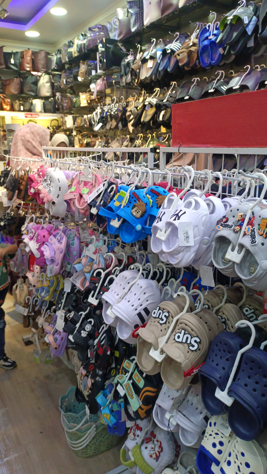 This image depicts the interior of a shoe store in Padang Panjang, Indonesia, likely a street-level shop given the informal display of goods. The store is densely packed with various footwear, primarily casual sandals and slippers, displayed on multiple racks and shelves. The merchandise includes children's shoes with cartoon characters like Mickey Mouse and other animated figures, as well as adult footwear such as clogs and flip-flops. Several pairs of shoes are hung with white plastic hangers on metal racks, creating a colorful and varied display. Some of the shoes are white with black accents, featuring logos like "NY." Others are brightly colored, with blue and tan options prominently visible. In the background, more shoes and handbags are visible on higher shelves. A person, partially visible on the left, is wearing a headscarf and a dark colored jacket. The lighting suggests an indoor setting with artificial lights overhead, some of which appear to be fluorescent. The overall impression is a busy, retail environment focused on affordable footwear.