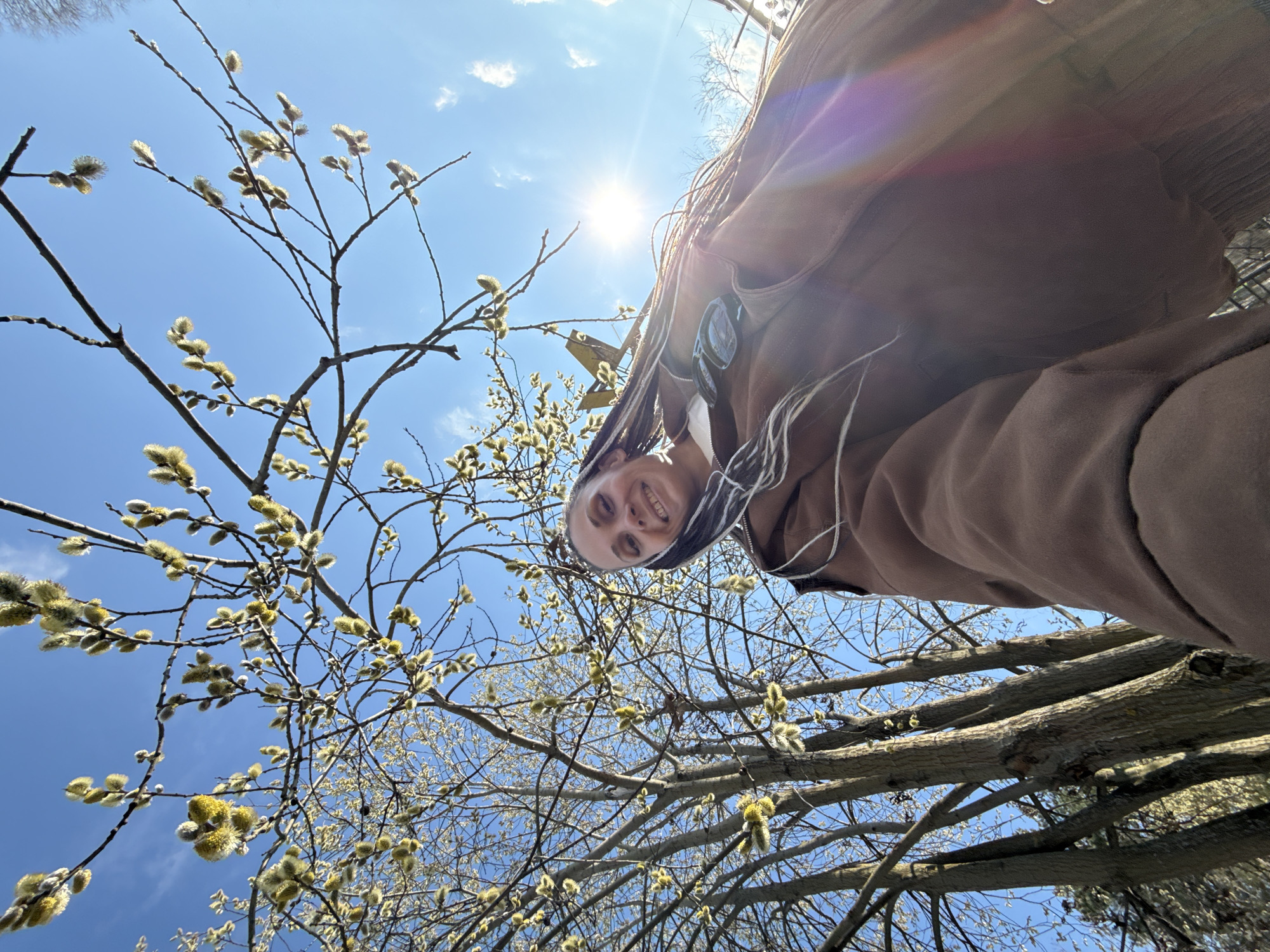 A low-angle, upward-facing photograph captures a woman smiling while looking down at the camera. She is partially obscured by the branches of a tree with fuzzy catkins, set against a bright blue sky with the sun shining brightly. The woman is wearing a brown hooded sweatshirt or jacket. The location is presumed to be the city of Rusanovo, Russia.