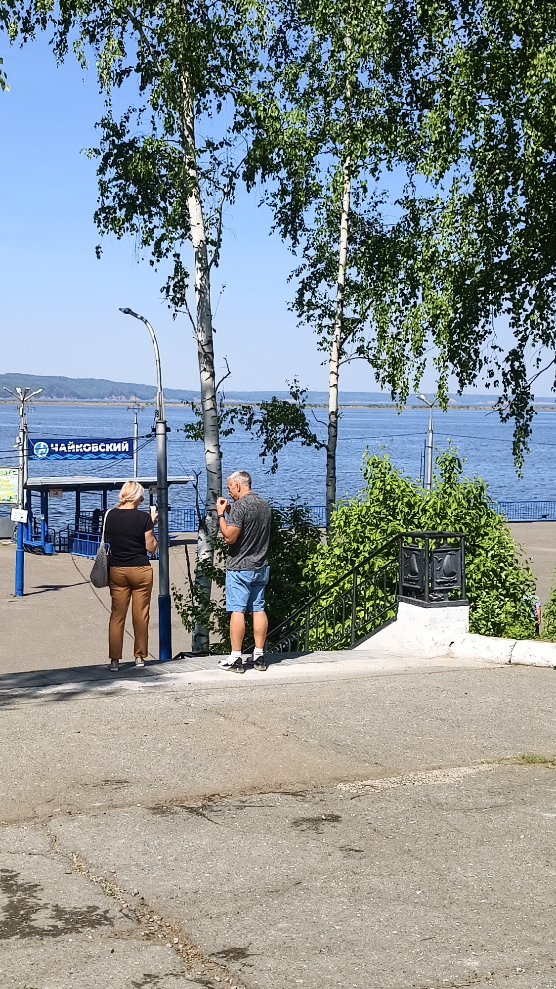 The image captures a scene in Chaikovsky, Russia, likely near a riverfront or lakeside. A man and a woman stand near a viewing point overlooking a body of water. The woman is on the left, wearing a black top and brown pants, and she appears to be using her cell phone. The man stands to the right, in a gray t-shirt and blue shorts, and seems to be putting something in his mouth, or smoking. They are situated on a concrete surface, with birch trees and greenery nearby. In the background is the body of water, with visible shoreline and vegetation on the opposite side. A sign with the word "Чайковский" (Chaikovsky) is visible in Cyrillic script, indicating the location. A set of black iron steps with a white wall and railing leads down to a lower level. The sky is clear and blue, suggesting a sunny day.