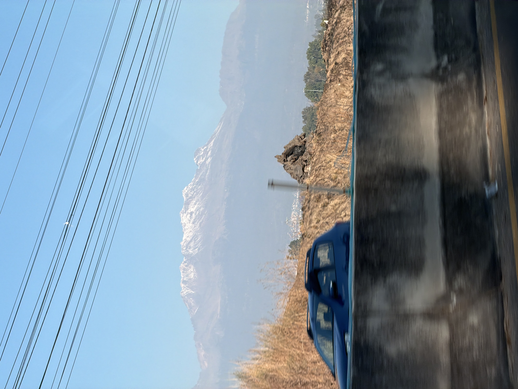 The image presents a daytime view captured from a moving vehicle in the vicinity of Santa María Zacatepec, Mexico. In the immediate foreground, a concrete highway barrier runs horizontally across the lower third of the frame, showing motion blur, with a yellow road line visible on the asphalt below. A dark blue hatchback vehicle, equipped with roof rails, is partially visible above the barrier in the left-center, also blurred due to motion. Behind the barrier, the terrain consists of dry, brown earth with scattered sparse vegetation, leading to a small, dark, irregular rock formation on the right side of the mid-ground. Further in the distance, green trees partially obscure a cluster of white and light-colored structures, indicative of a town or city, with a prominent street light pole visible. The background is dominated by a large mountain range with snow-capped peaks, partially veiled by atmospheric haze. The sky above is clear blue, and a faint crescent moon is discernible in the upper right quadrant. Multiple parallel black utility lines traverse the upper portion of the sky diagonally.