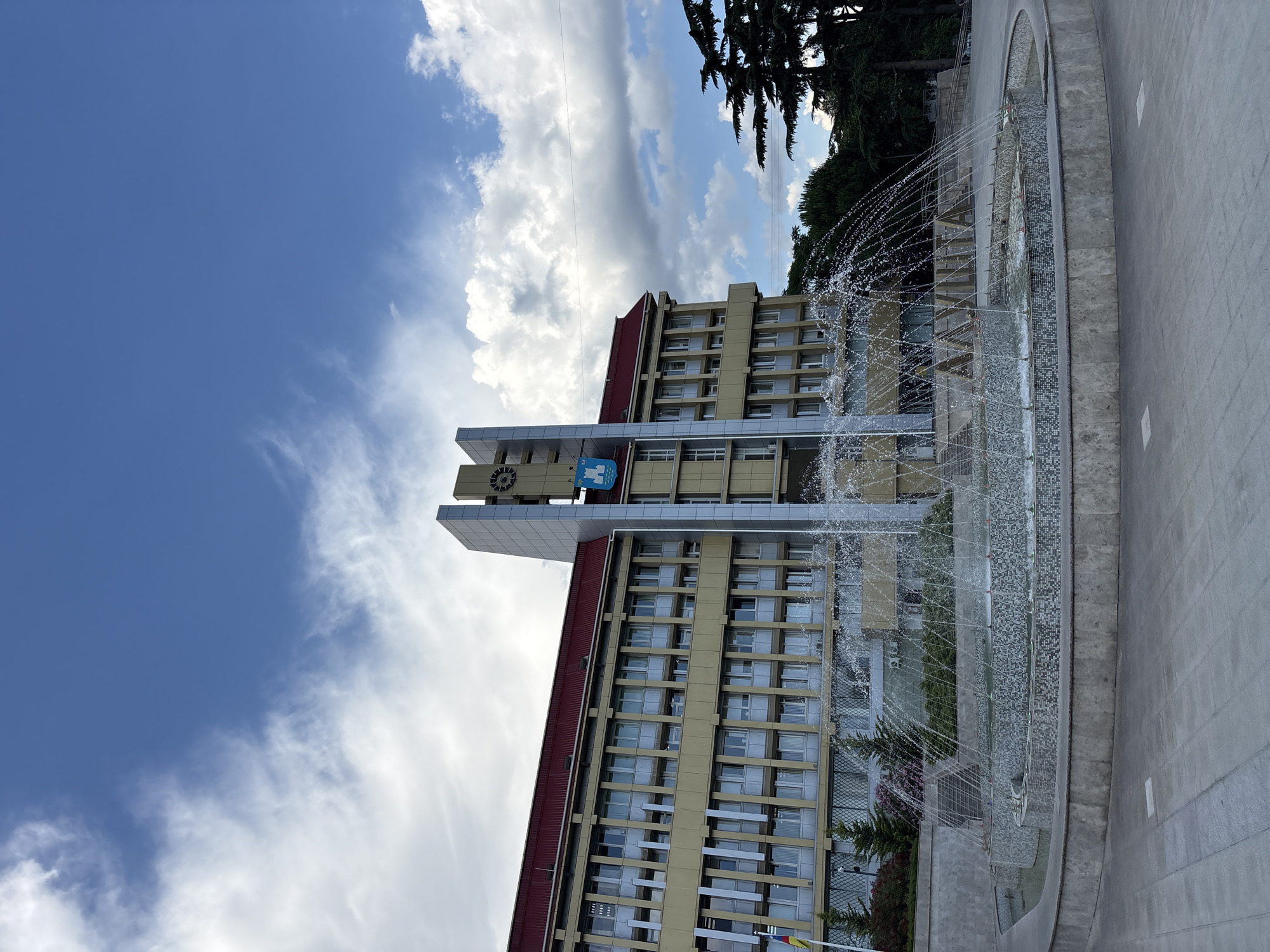 In the city of Alushta, Ukraine, a multi-story building with a red roof stands behind a circular fountain. The building is a pale yellow color and has many windows. A clock tower is attached to the front of the building, and a blue emblem is visible on it. The fountain is in the foreground, with water spraying outwards in arcs. The area around the fountain is paved with grey tiles. The sky above is blue with some white clouds. There are trees to the right of the building, and some greenery in front of it. There are no people visible in the image.