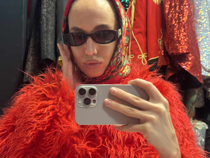 A person is depicted in a chest-up selfie taken within an interior space in Dubai, United Arab Emirates. The subject wears dark oval sunglasses, a patterned headscarf (balaclava-style) primarily featuring red, green, and gold floral motifs, and a voluminous, bright red shaggy faux fur-like garment. Their left hand is raised, with fingers touching their temple, revealing a ring on the ring finger. A light gray smartphone, equipped with a triple-lens camera array, is held horizontally in the subject's right hand, actively capturing the image. The background consists of various hanging garments on racks, including a dark, possibly textured piece on the left, a red garment with gold trim in the center, and a highly reflective, bright red sequined garment to the right, indicative of a retail clothing store or costume shop environment.