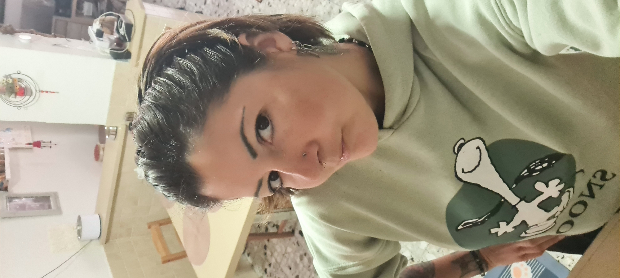 A selfie captures a woman indoors, wearing a light green hooded sweatshirt featuring a Snoopy graphic. She has dark hair, pierced ears, and nose piercings on both sides of her nose, with visible dark eyebrows. The background includes a table with a light-colored top, a white wall with various decorations, including a picture frame and a hanging ornament. A motorcycle helmet is also visible on a shelf in the background. The location is not identifiable as Nahariyya, Israel, based on visual information alone.