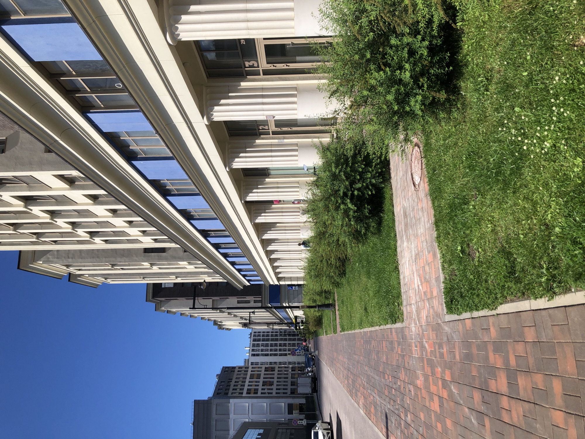The image depicts a street scene in Saint Petersburg, Russia, on a clear, sunny day. The perspective is from a paved sidewalk, looking down the street towards the right. The sidewalk is made of red and brown interlocking bricks. To the right of the sidewalk is a strip of green grass with several mature, leafy bushes.
Dominating the right side of the image is a modern, multi-story building with a light-colored facade and a band of blue panels above a row of windows. This building features a colonnade of tall, fluted columns at ground level, supporting the upper structure. Two doorways are visible, marked with the numbers "2" and "3", and one has the number "29" above it.
Further down the street on the right, other modern buildings can be seen. The left side of the street shows a road with several cars, including a white sedan and a white truck. In the distance, more buildings of varying architectural styles are visible, suggesting an urban environment. The sky is a clear, bright blue, indicative of good weather. A single person, appearing as a dark silhouette, is standing near the building on the right, partially obscured by bushes. There are no other discernible human interactions or specific actions taking place. The environment appears clean and well-maintained, typical of an urban setting.