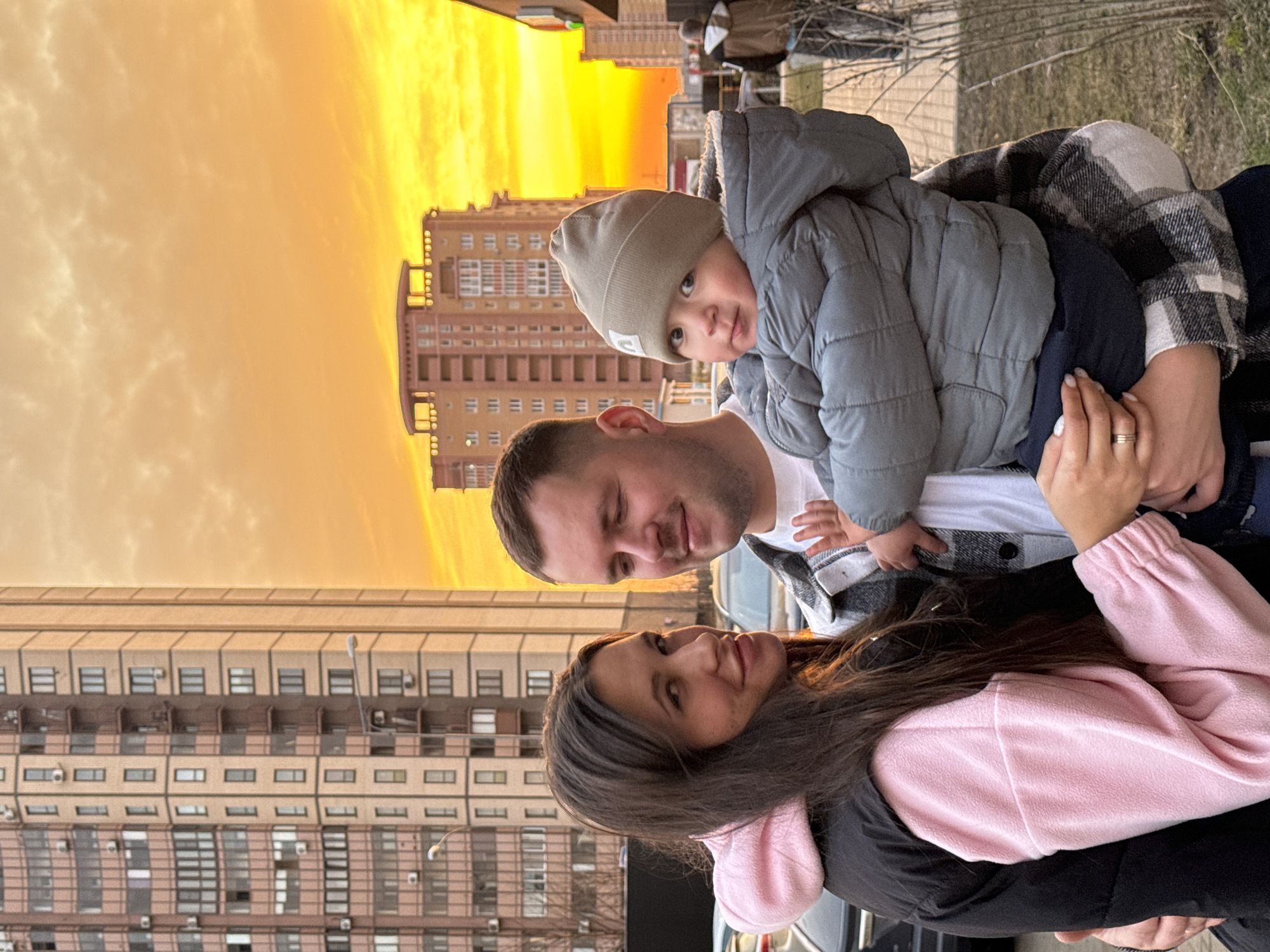 A daytime outdoor portrait captures three individuals: an adult female, an adult male, and an infant, posing against a backdrop of urban residential buildings under a vivid orange and yellow sky, indicative of sunset or sunrise.
The adult female, positioned on the left, has long dark hair and wears a pink fleece hoodie under a black puffer vest. She looks towards the camera with a subtle smile, her left arm partially visible around the male adult's lower back.
The adult male, centrally located, has short brown hair and wears a white t-shirt beneath a black and white plaid flannel shirt. He holds the infant in his arms, his gaze directed towards the infant with a slight smile. A gold wedding band is visible on his left ring finger.
The infant, held by the male adult, is dressed in a grey quilted winter jacket with a fuzzy lining and a matching grey knit beanie. The infant looks directly at the camera with a neutral expression.
The background features multiple tall, multi-story residential apartment buildings, characterized by numerous windows and balconies. These structures are typical of modern and late Soviet-era residential districts in Moscow, Russia, such as those found in areas like Yasenevo or Chertanovo. A streetlight is visible on the left, and partially obscured parked vehicles are present. The immediate foreground consists of sparse dry grass and leafless bushes on a paved surface. A blurred figure of another person is visible in the far right background.