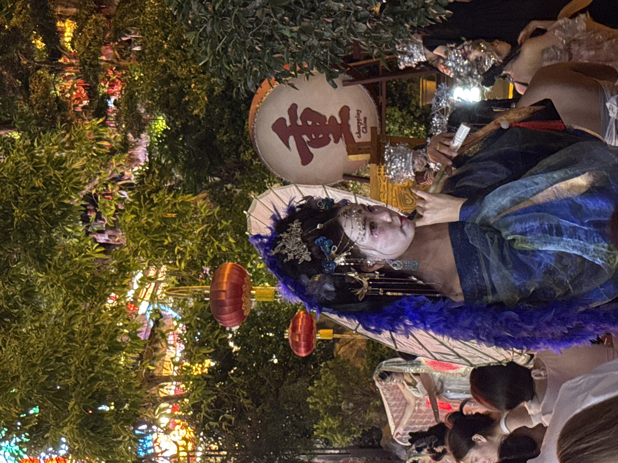 This image depicts a night scene with a performer dressed in traditional Chinese attire, including a black wig with blue feathers and elaborate headdress, white face paint, and a flowing blue and green robe. The performer is holding a long, hollow instrument to their mouth, suggesting they are playing music. In the background, a large drum with a red Chinese character "重" (Chóng) and the text "Chongqing China" is visible. Red lanterns hang from trees adorned with colorful lights, creating a festive atmosphere. Several other individuals, also in costume with ornate headdresses, are present, some partially visible. The setting appears to be an outdoor event or festival in Chongqing, China.