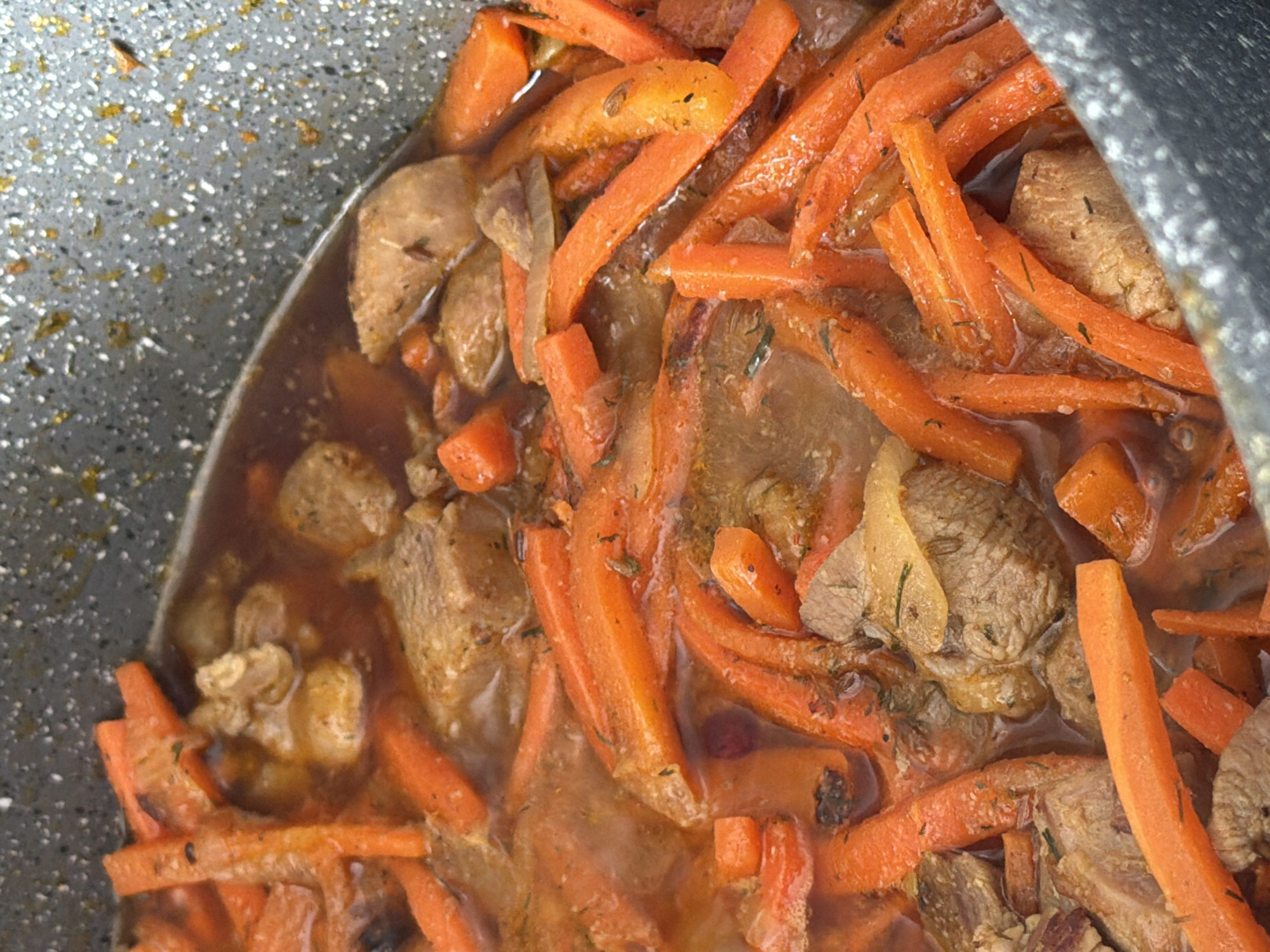 A close-up, overhead shot shows a thick, dark stew in a speckled grey pot, likely a Dutch oven or similar heavy-bottomed pot. The stew contains large, irregular chunks of braised meat, appearing to be beef, alongside vibrant orange carrot sticks and sliced onions. The carrots are cut into lengths resembling julienne, though some are thicker. The broth is a rich, reddish-brown color, suggesting a long simmering time. Small green herbs, possibly dill or parsley, are sprinkled throughout the dish. The pot's rim is visible along the top and right edges of the frame. The overall impression is one of a hearty, slow-cooked meal. The location is identified as Водоканал Снт, Russia, implying this is likely a home kitchen or outdoor cooking environment within a dacha or garden cooperative. There are no people or distinct geographical features visible.