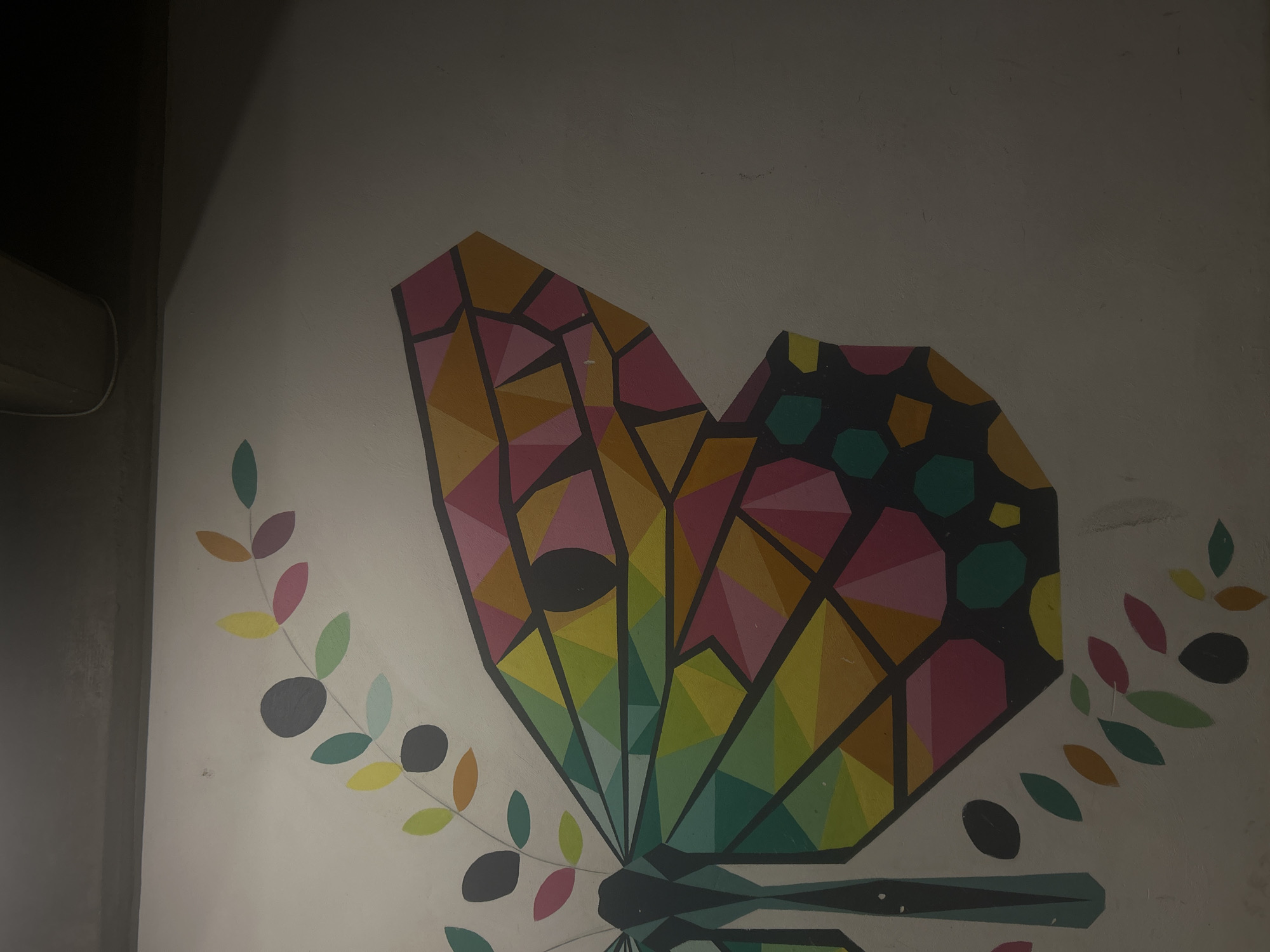 The image shows a mural painted on a white wall. The mural depicts a stylized butterfly with geometric shapes and a vibrant color palette, including pink, yellow, green, and teal. The butterfly's wings are bordered by stylized branches with leaves in similar colors. The location is not specified, so the city and area of "Unknown, Unknown" cannot be confirmed.