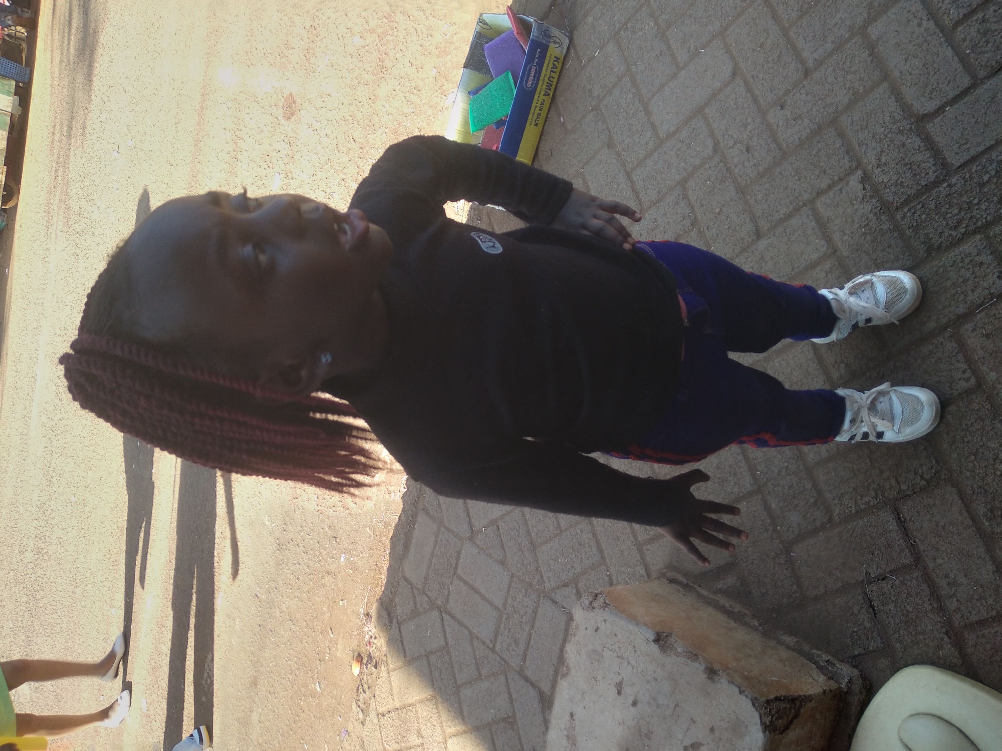 The image depicts a young Black girl with braided hair, wearing a black long-sleeved shirt and blue pants with red stripes. She is standing on a paved surface with some shadows cast on it. To the right of the girl, there is a cardboard box containing several colorful sponges, predominantly green, purple, and red. In the background, a portion of a green and white vehicle is visible. The overall environment appears to be an outdoor setting, possibly a street or market area, in Nyeri, Kenya, as suggested by the presence of local merchandise and the style of the paving.