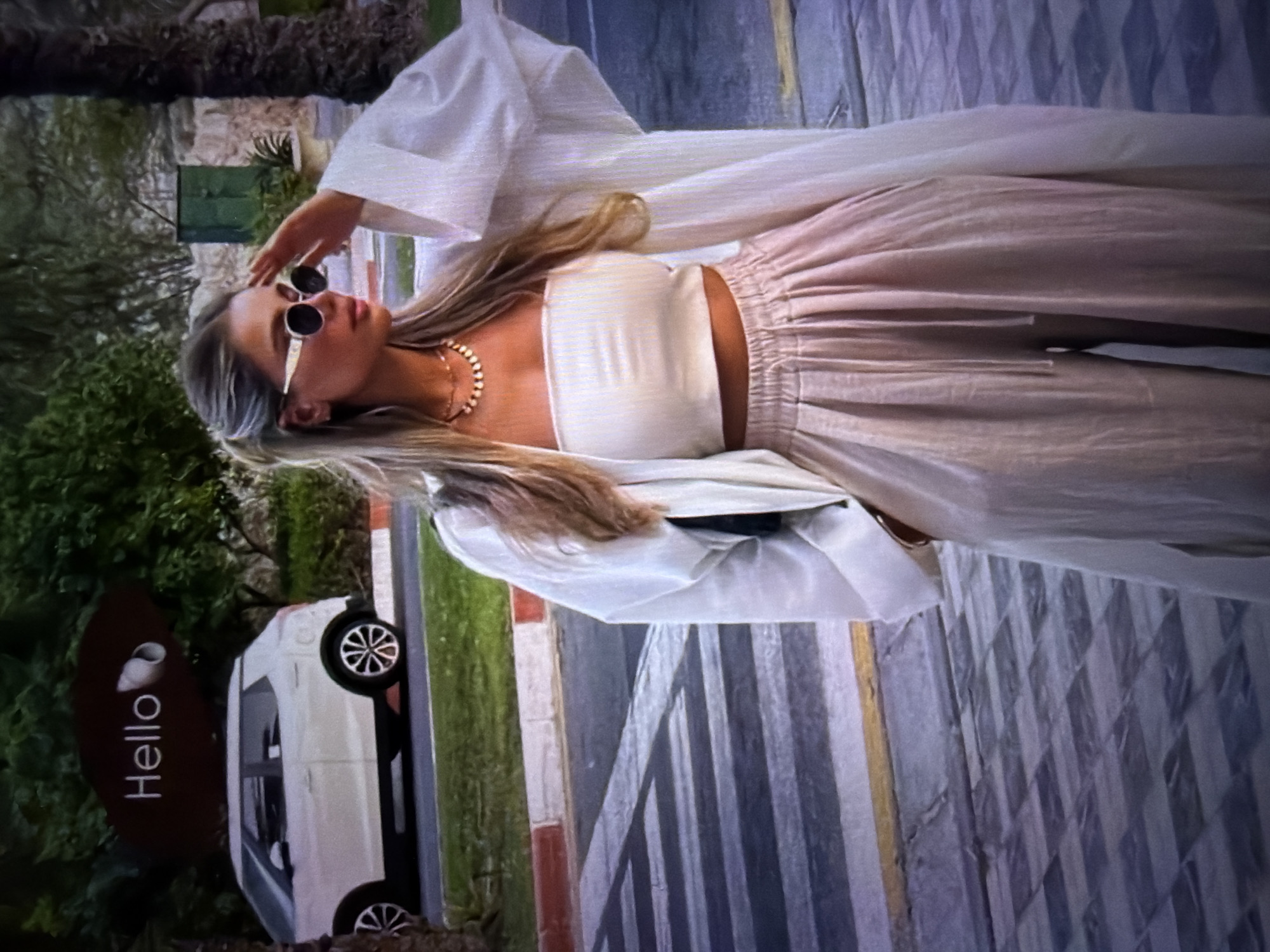 The image shows a woman posing outdoors. She has long blonde hair and is wearing sunglasses, a white crop top, a white duster, and flowy pink pants. She is also wearing a gold necklace. The woman is standing in front of a white van and a sign that says "Hello". The background includes greenery, a street with crosswalk markings, and a building. The lighting suggests it is daytime. The overall mood is relaxed and stylish. The image is oriented vertically.